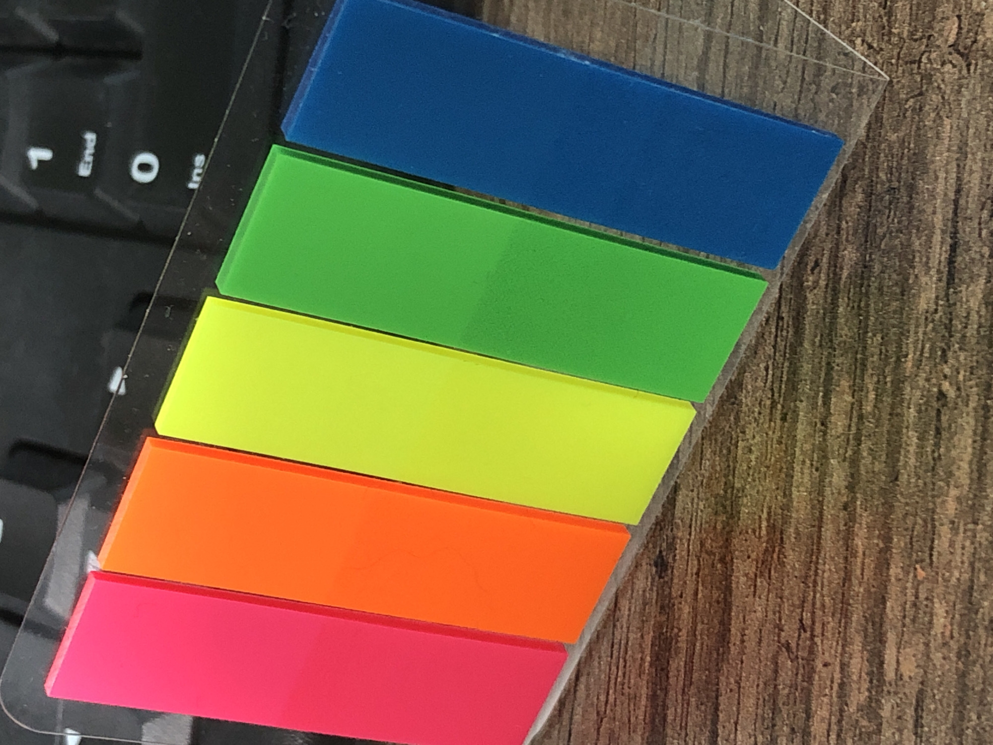 The image shows a close-up, overhead view of five brightly colored sticky notes, arranged diagonally on a wooden surface. The sticky notes are, from left to right: neon pink, orange, neon yellow, bright green, and blue. The sticky notes appear to be on a transparent backing, possibly part of a dispenser. Behind the sticky notes, a portion of a black keyboard is visible. The wooden surface has a prominent grain pattern. The lighting is from above and to the left, casting subtle shadows. There are no people or animals present. The location of Petropavlovsk-Kamchatsky, Russia is not discernible from the image.