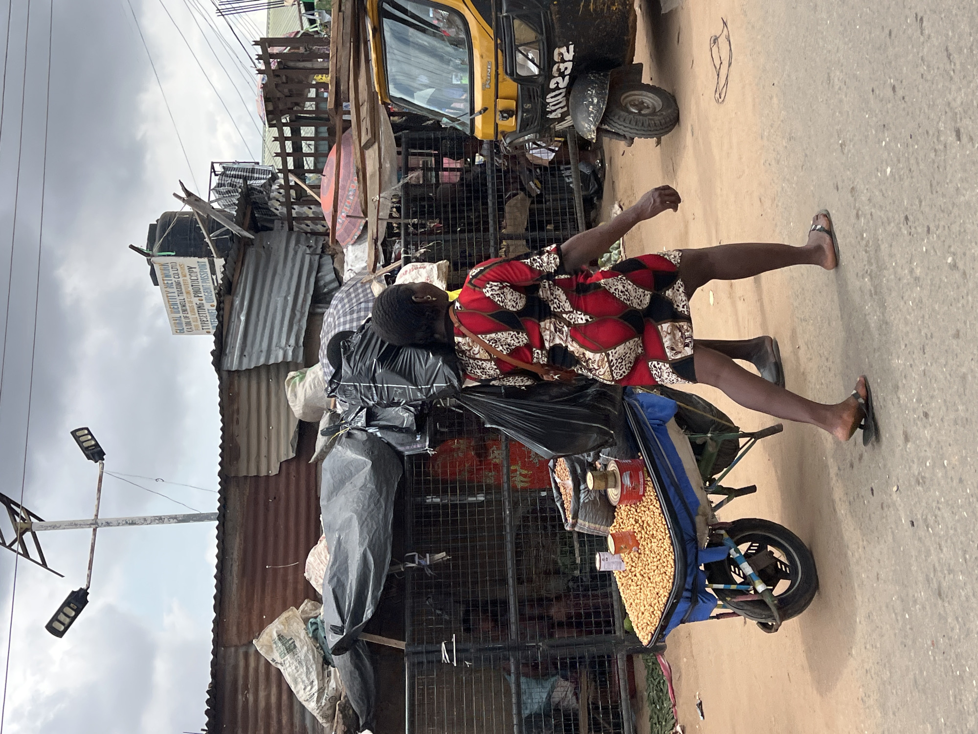 This image captures a vibrant street scene in Nkpor, Nigeria. A woman, dressed in a red, black, and white patterned dress, walks along a dusty road. She is carrying a large black bag and has a smaller shoulder bag. Next to her, a blue cart is filled with what appears to be peanuts. The cart is attached to a yellow tricycle. In the background, various structures made of corrugated metal and wood line the street, suggesting a bustling marketplace or informal settlement. A sign for "GLOBAL IDENTITY NETWORK" with services like "TYPESETTING PHOTOCOPY" and "INTERNET URGENT PASSPORT" is visible on one of the buildings. The sky is overcast, indicating a cloudy day. The overall impression is one of daily life and commerce taking place in a lively urban environment.