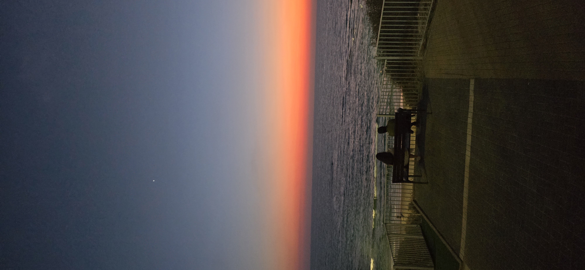 A tranquil scene unfolds at dusk in Bat Yam, Israel, with a couple silhouetted on a bench, facing a vast, calm ocean. The sky transitions from a deep, dusky blue at the top to vibrant shades of orange and red near the horizon, indicating the sun has recently set. A single star is visible in the upper left quadrant of the sky. The ocean stretches out, its surface rippled by gentle waves. In the foreground, a paved area with a subtle pattern is visible, bordered by a white railing and green landscaping. The overall atmosphere is one of peaceful contemplation.