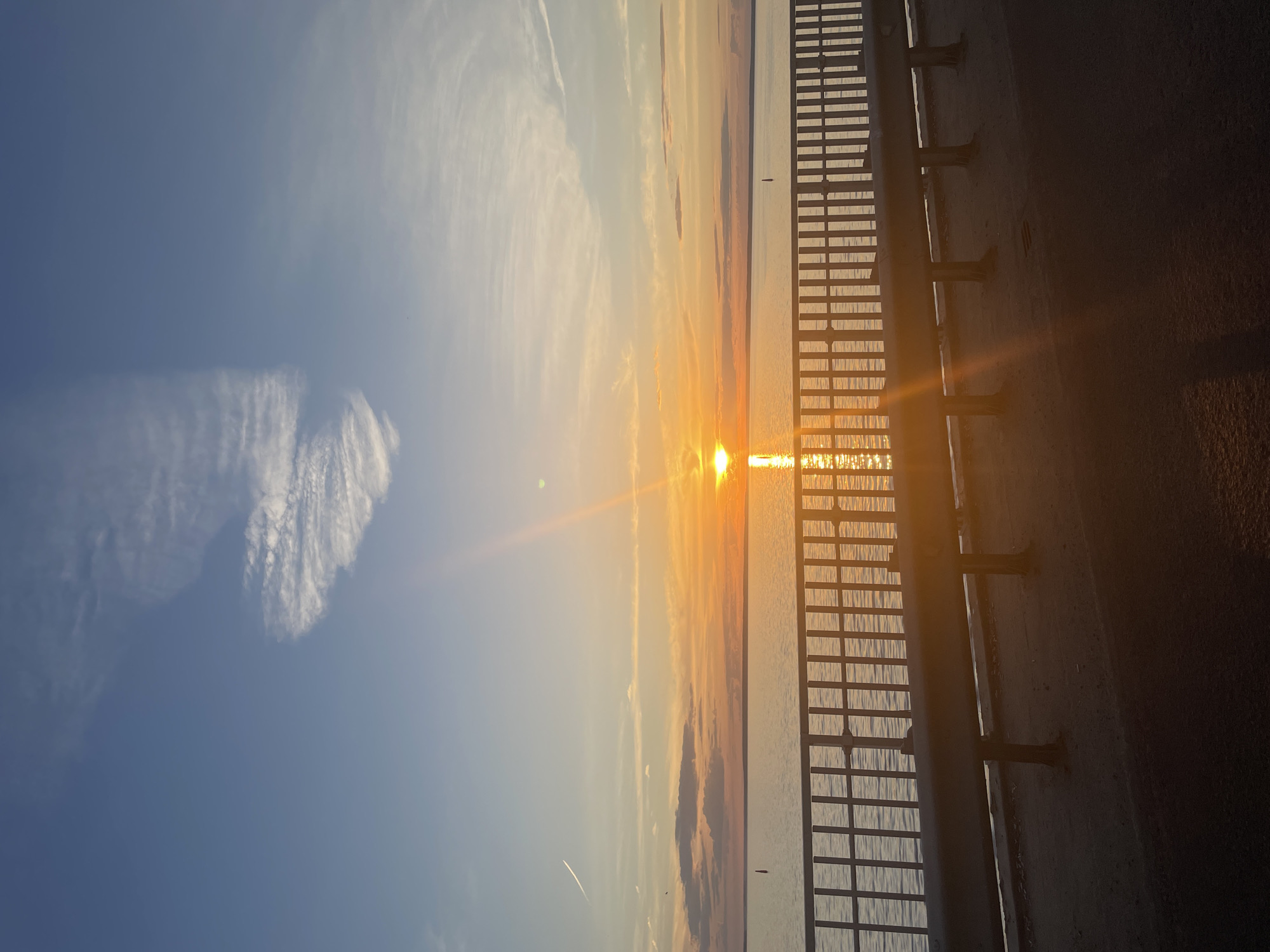 A wide-angle, vertical shot captures a sunset over a body of water, likely a lake or ocean, with a bridge in the foreground. The sun, a bright orange orb, is positioned just above the horizon, casting a shimmering reflection on the water's surface. The sky transitions from a pale blue at the top to warm hues of orange and yellow near the horizon, dotted with wispy clouds. A metal railing runs horizontally across the middle of the frame, separating the viewer from the water. The railing is supported by metal posts, and a guardrail runs along the top. The ground in front of the railing appears to be a paved surface. The overall scene evokes a sense of tranquility and natural beauty.