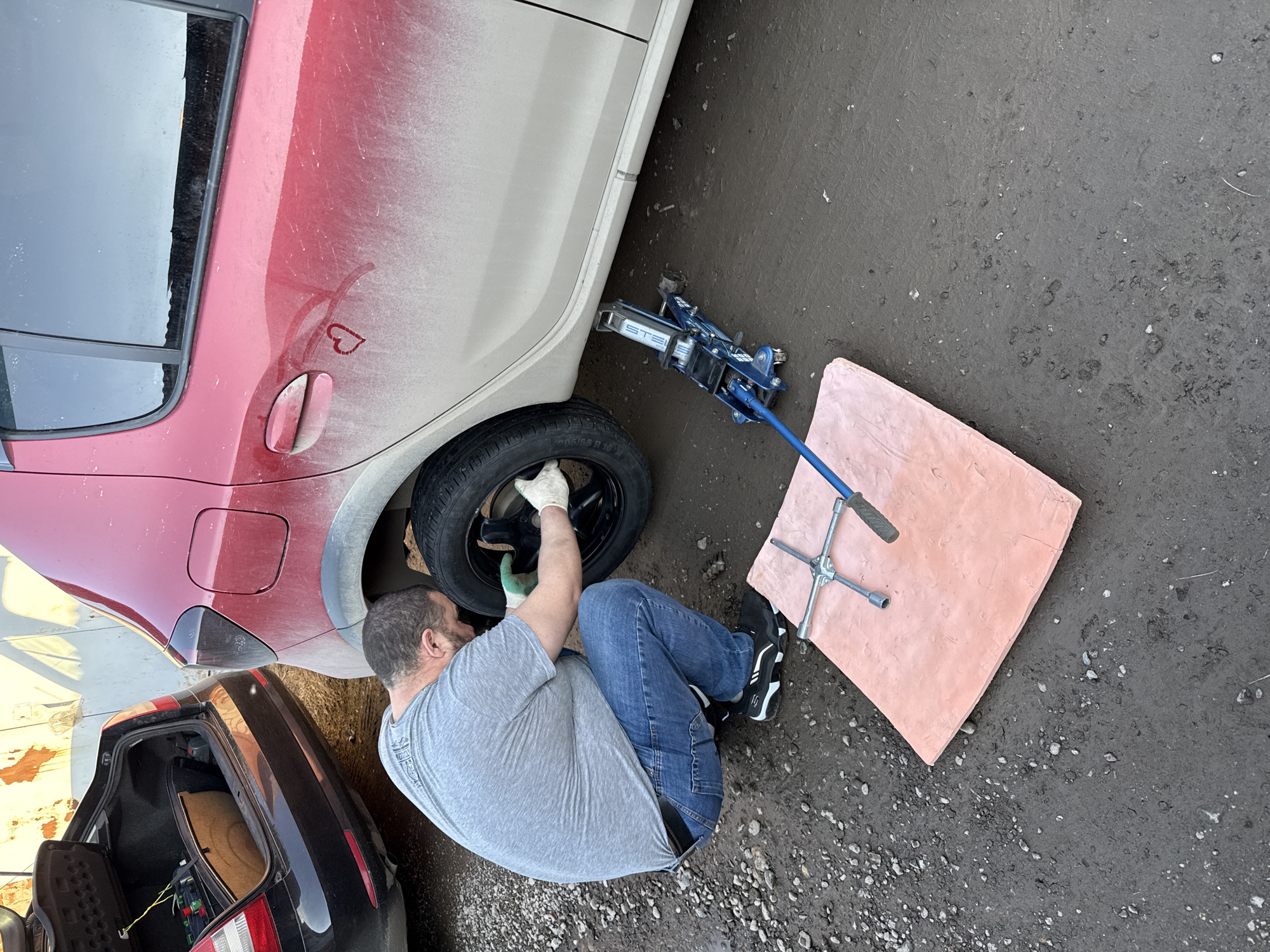 A man is changing a car tire outdoors, likely in Izhevsk, Russia. He is crouching next to the car, wearing a grey t-shirt, blue jeans, and black and white sneakers, and is working on the tire with gloved hands. The car is two-toned, with a red rear panel that is dirty. A blue hydraulic jack is supporting the car. A lug wrench rests on a pink square mat nearby. The ground is paved and dirty. The open trunk of a dark-colored car is visible in the lower left corner of the image. A heart is drawn in the dirt on the rear panel of the red car.