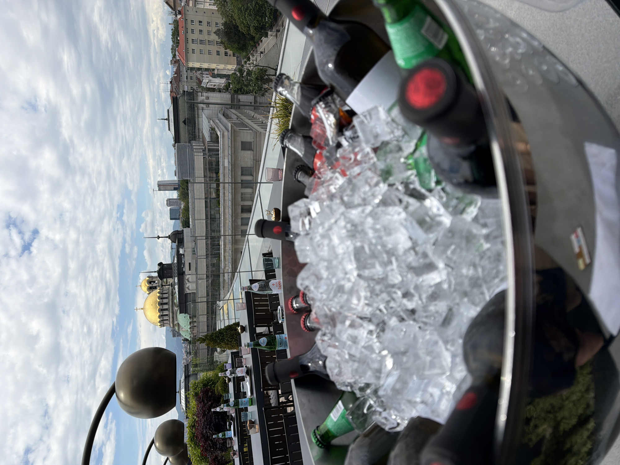 The image shows a rooftop view from Sofia, Bulgaria, with the gold dome of the Alexander Nevsky Cathedral visible in the background. A silver ice bucket filled with ice and bottles of wine and sparkling water occupies the foreground. A table and chairs are set for service near the edge of the rooftop. In the distance, buildings and the skyline of Sofia are visible. The sky is cloudy. Large black spherical sculptures are also visible near the rooftop edge.