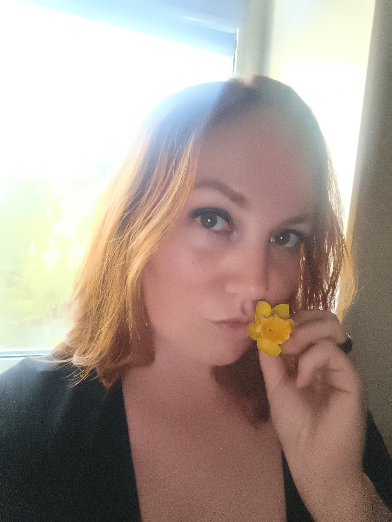 The media file is a close-up, self-portrait of a woman with reddish-blonde hair. She is holding a small yellow flower near her lips, as if about to kiss it. Her eyes are open and looking directly at the camera, and her lips are pursed. She is wearing a dark top, and a ring is visible on her right hand. The background is bright, suggesting it is daytime and possibly sunny, with a window or light source behind her. The overall mood appears gentle and perhaps a little playful, with the woman's expression being soft. The location context provided is Chaikovsky, Russia, but there are no specific visual cues in the image to confirm this. There is no visible text in the image.