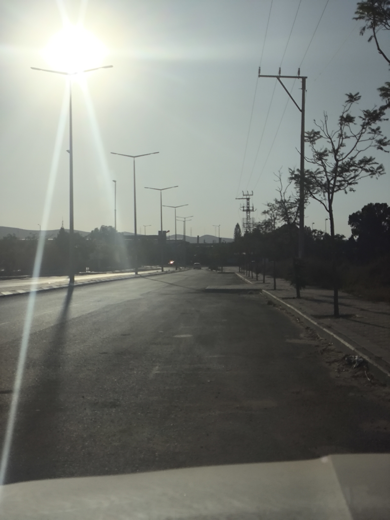 The image shows a road with streetlights and power lines under a bright, hazy sky, likely during the day. The perspective appears to be from inside a vehicle. The road is asphalt and has white markings. On the left side of the road, there are several tall streetlights, with one in the foreground casting a long shadow. On the right side, there are trees and more power lines. In the distance, there are hills and buildings. The scene is consistent with a road in or near the city of Dimona, Israel, given the presence of characteristic infrastructure and landscape features of the Negev region. There are no visible people or specific actions taking place, and the overall impression is one of a somewhat desolate, semi-urban or suburban environment.