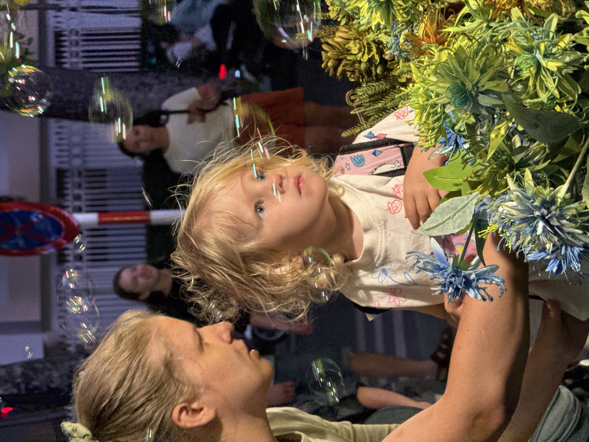 Captured in a vertical orientation, the image portrays a tender moment between an adult and a young child in an urban setting at night, likely in Nha Trang Ward, Vietnam.
In the foreground, an adult, possibly a woman with blonde or light brown hair, is visible on the lower left, looking upwards. Her arms are holding up a young, blonde-haired child, who is the central focus of the image. The child, dressed in a light-colored t-shirt adorned with cartoon patterns and a pink strap (possibly from a bag or garment), gazes upward with wide-eyed curiosity and wonder.
Numerous iridescent soap bubbles, some round and some elongated by movement, float around the child, reflecting vibrant colors from the ambient lights. To the upper right, a large cluster of artificial green and blue flowers and foliage fills part of the frame, adding a decorative element.
The background is dark and softly blurred, indicating night or late evening. Reflections of lights and indistinct urban structures are visible, suggesting a city street or public space. Several other blurred figures of people can be seen in the background, including a man in a white shirt further back. On the left, a red and white striped barrier and a partially visible blue circular traffic sign with a white 'X' and a red diagonal line are discernible.
The scene depicts an engaging activity where the adult is sharing the simple joy of bubbles with the child, whose expression radiates innocent delight. The glowing bubbles against the dark background create a magical and heartwarming atmosphere.