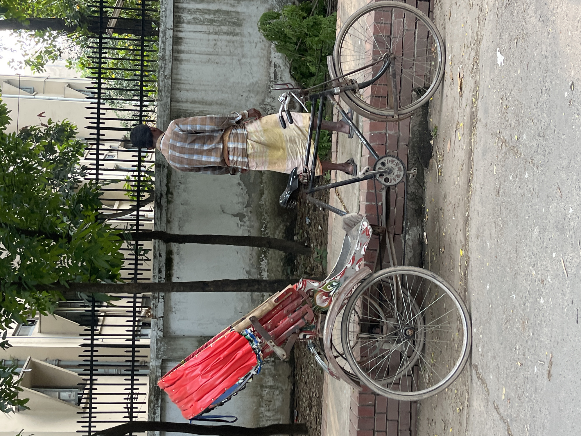 A man is standing next to a rickshaw on a paved street in Dhaka, Bangladesh. The man is wearing a plaid shirt and a yellow lungi. The rickshaw has a red canopy and ornate details. A wrought iron fence and building are in the background. Trees frame the scene.