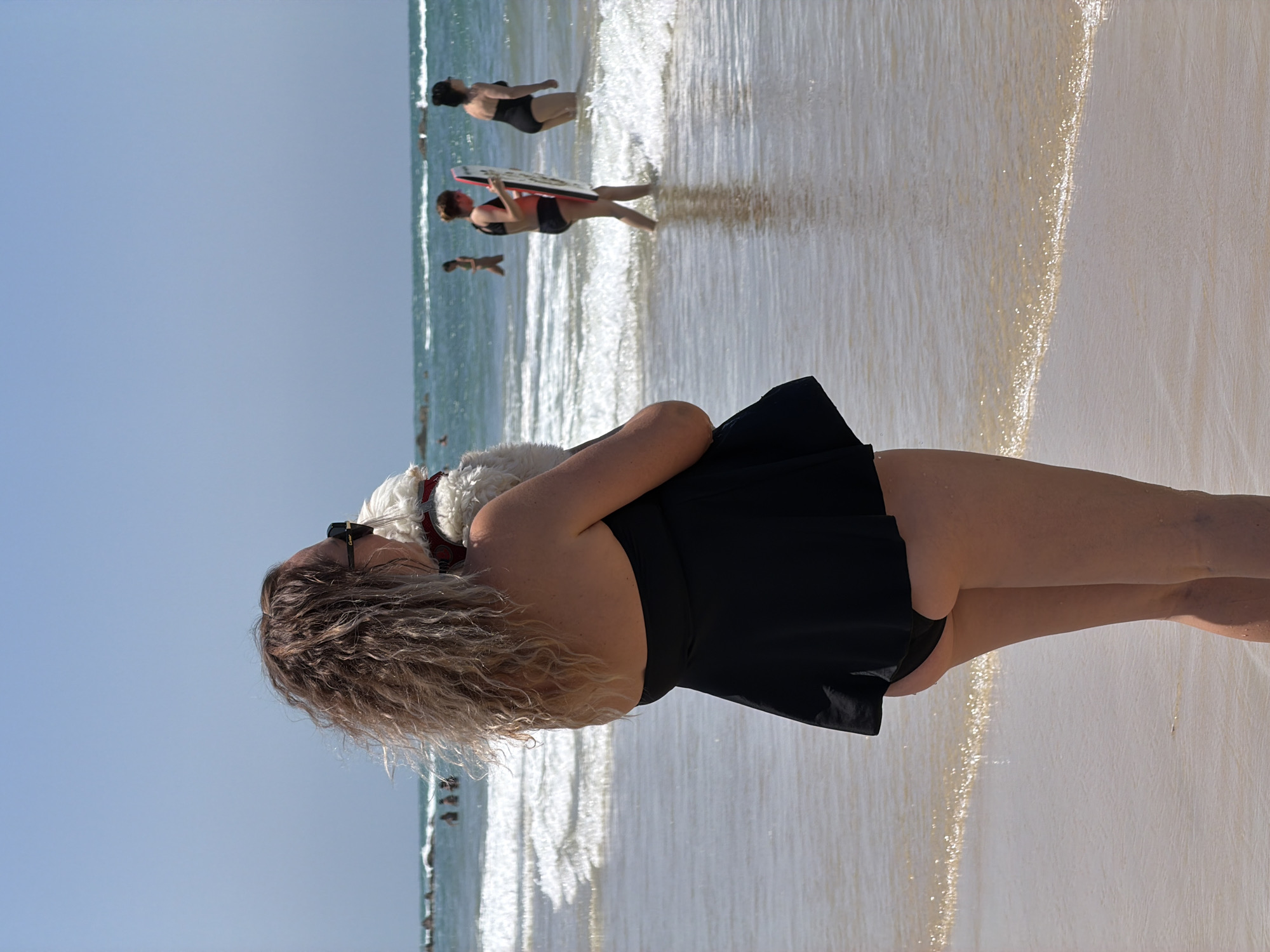 On a sunny day at a beach in Bat Yam, Israel, a woman with curly blonde hair and sunglasses stands facing the water. She wears a black swimsuit and cradles a small white dog in her arms. Behind her, several people are in the ocean, some swimming and some with boogie boards. The beach has wet sand near the water's edge, transitioning to drier sand further back. The sky is clear and blue.
