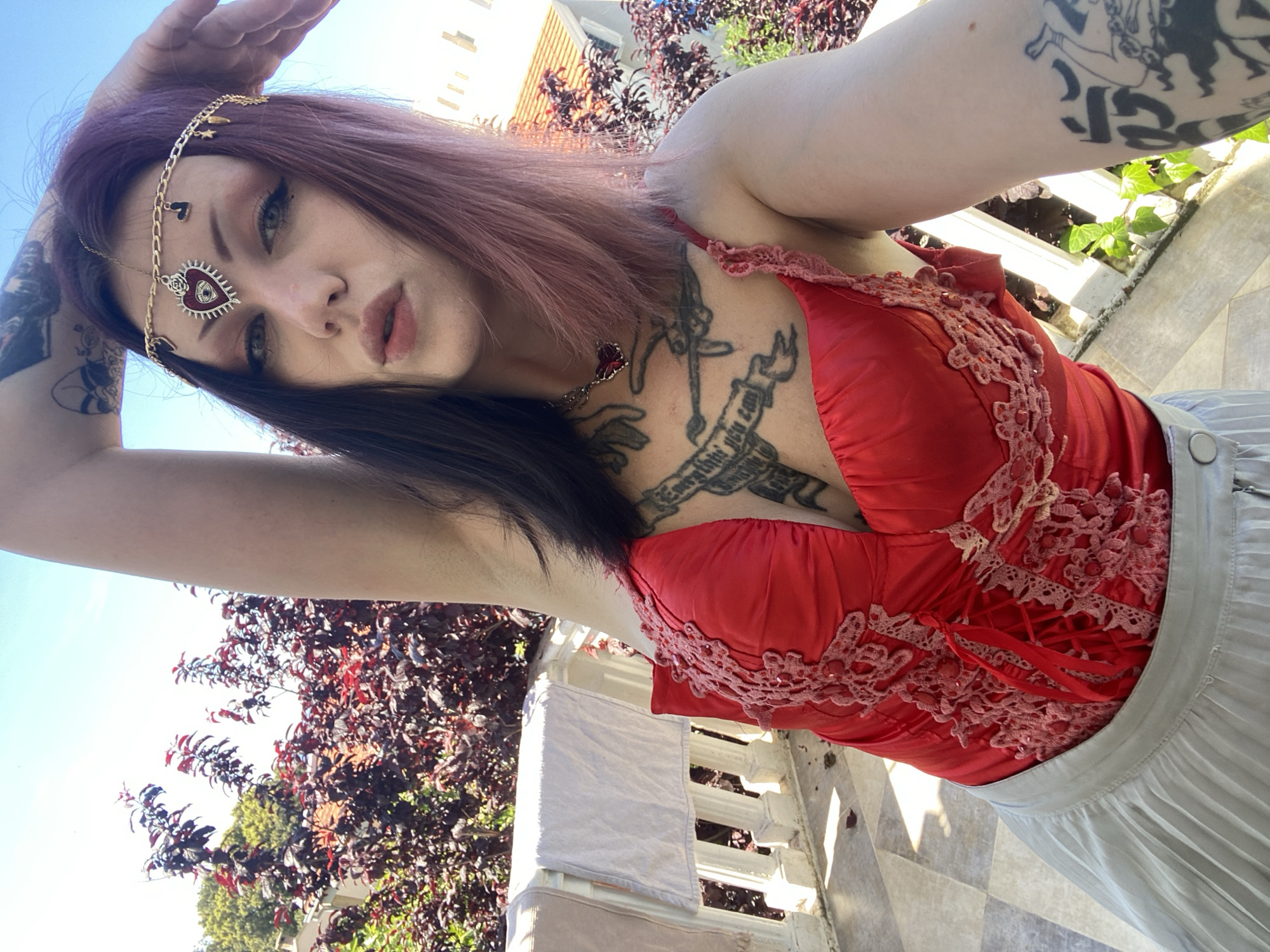 A young woman with long, dark roots blending into purple-pink hair poses for a selfie in bright daylight. Her gaze is direct, accentuated by prominent dark eyeliner and light-colored eyes, and she has a philtrum piercing. She wears a golden headpiece adorned with small charms like a heart, stars, and a crescent moon, culminating in a central red heart-shaped evil eye pendant on her forehead. A matching red heart pendant hangs from her neck.
She is dressed in a vibrant red top, possibly a corset, featuring intricate light pink lace and beadwork on the front and visible lacing at the back. A light grey pleated skirt completes her outfit. Her left arm is covered in a dark, intricate tattoo sleeve depicting figures and patterns, while her chest features additional tattoos, including a banner that partially reads "EVERYTHING YOU CAN". Her right arm is raised, partially obscuring the top of her head as she takes the photo.
The scene is an outdoor setting, likely a balcony or terrace in Kunje, Montenegro, under a clear blue sky. Lush foliage with dark reddish-purple and green leaves forms a natural backdrop. A white balustrade or railing and a light-colored tiled floor are visible to her right. In the upper background, a light-colored building with an orange-tiled roof can be seen. The bright sunlight indicates it is daytime, casting clear shadows.