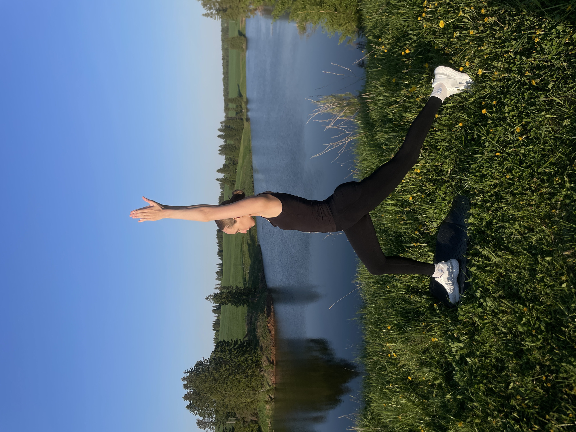 A woman is performing a yoga pose in a grassy area next to a body of water. She is wearing black athletic clothing and white sneakers. She is in a Warrior III pose with her arms extended forward and one leg extended behind her. The water reflects the sky and trees on the opposite bank. The grass is green and has small yellow flowers. The location is Подшивалово, Russia.