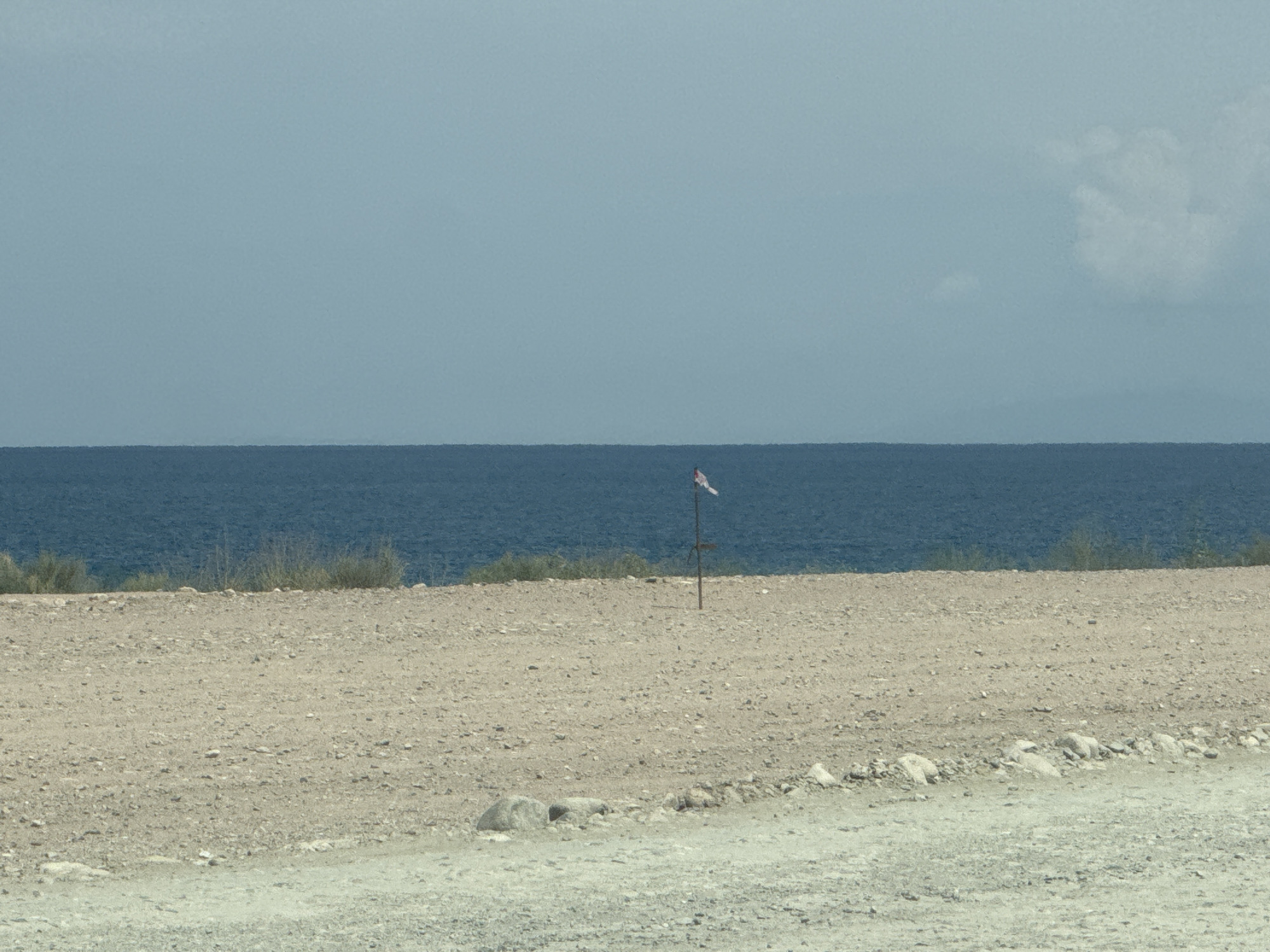 The image shows a desolate, arid landscape overlooking a body of dark blue water. In the foreground, a rough, gravelly dirt path leads up to a barren, sandy expanse. Scattered rocks are visible on the ground. A thin, weathered wooden pole stands in the middle ground, topped with a small, striped flag. Behind the pole, a line of sparse, dry vegetation marks the transition to the water. The water extends to the horizon, meeting a pale blue sky that has a few wispy clouds on the right side. The scene suggests a remote, possibly coastal or lakeside location. There is no clear indication of urban infrastructure or recognizable landmarks of Tong, Kyrgyzstan.