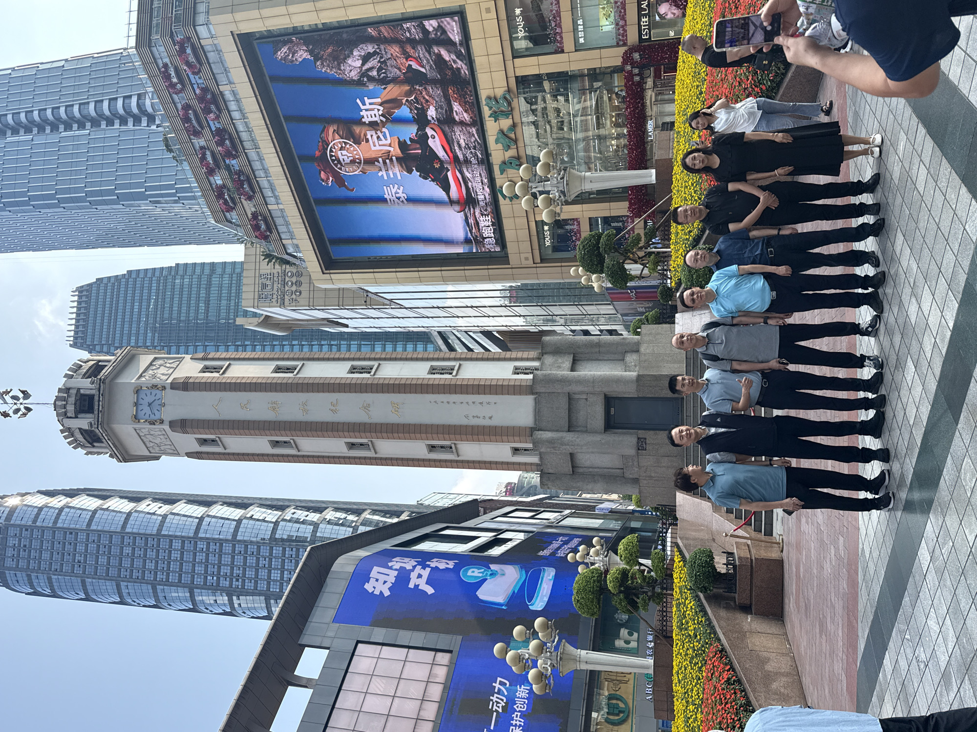 The image, depicting a sunny day in Yuzhong District, China, captures an urban street scene where a group of people is posing for a photograph. In the foreground, ten individuals, consisting of men and one woman, are arranged in a neat line on a paved sidewalk, looking towards the camera. Many are dressed in semi-casual attire, some smiling. A person's hands and a smartphone are visible in the upper right, indicating they are taking the group's picture.
Behind the posing group, a vibrant flower bed with yellow and red blooms borders the pathway, separating it from the commercial buildings. A distinctive, light-colored clock tower with multiple windows and Chinese characters inscribed on its facade stands prominently in the mid-ground. Several modern high-rise buildings with glass exteriors rise into a clear sky, dominating the background.
Large digital billboards are a key feature of the commercial environment. One prominent screen advertises "泰兰尼斯" (Tailannisi) running shoes, showing a person in sportswear and the tagline "稳跑鞋 更懂校园运动保护" (Stable running shoes, better understanding campus sports protection). Another blue billboard displays text such as "保护创新" (Protect Innovation) and "一动力" (One Power/Motivation). Visible storefront signs include "YOUS*", "滿記甜品" (Honeymoon Dessert), "ESTÉE LAUDER", "LAMER", and "ABC农业银行" (ABC Agricultural Bank of China). The scene combines modern city architecture with a touch of historical design in the clock tower.
