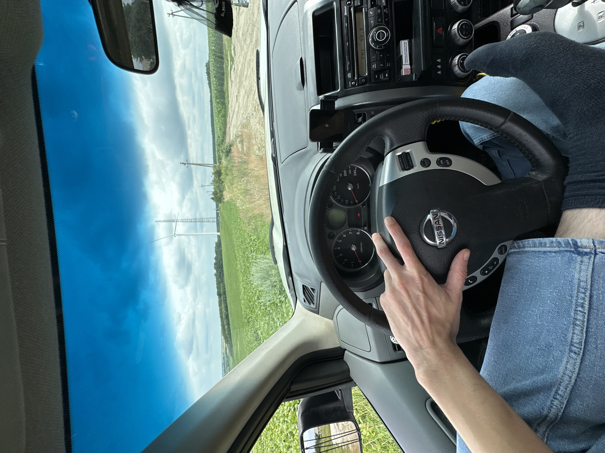 A first-person perspective from inside a Nissan X-Trail is captured as the vehicle navigates a dirt road through a rural landscape near Korchivka, Ukraine. The driver's left hand rests on the steering wheel, clad in blue denim jeans. A dark gray sock covers the right foot, which is visible near the center console. The dashboard displays a charging phone mount, a car radio, and climate control knobs. Outside, a vast field of green crops stretches towards a distant horizon, punctuated by power lines and a wind turbine. The sky above is a vivid blue, dotted with cumulus clouds, suggesting fair weather. The road appears unpaved and dusty, indicating the vehicle is likely off the main highway.