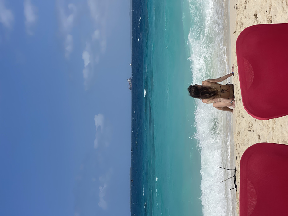 The scene depicts a beach in Zanzibar, Tanzania, under a clear blue sky with scattered white clouds. In the immediate foreground, the backs of two red beach chairs are partially visible, framing the view. A single individual, identified as female, is seated on the light-colored sand, facing away from the camera and looking out towards the ocean. She is wearing a dark bikini top, and her long, dark hair is visible. A white wave is breaking directly in front of her, and small pieces of driftwood or debris are present on the sand to her left. The ocean extends beyond the beach, exhibiting a clear gradient from turquoise near the shore to a darker blue further out. On the distant horizon, a large cargo vessel is visible, along with a small white sailboat to its right. A low, distant landmass or island is discernible on the far left horizon. No direct interactions between individuals are depicted.
