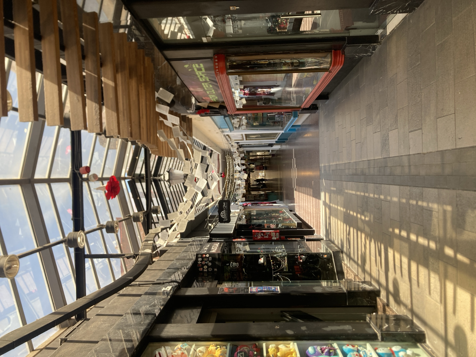 An interior view showcases a long pedestrian walkway lined with shops, bathed in sunlight. The shops are displayed on both sides of the corridor, each featuring glass-fronted display cases. The walkway extends into the distance, appearing narrower as it recedes. The perspective is skewed due to the tilt of the camera, which shows the architectural details of the upper structure of the walkway on the left.
The ceiling of the walkway is constructed of steel beams and glass panels, allowing abundant natural light to flood the space. Wooden slats hang decoratively from the ceiling, parallel to the shops. Attached to the wooden slats are numerous small, white paper pieces, creating a floating effect above the walkway. The floor is paved with light-colored stone tiles.
The shops' signage and merchandise are visible, indicating retail activity. The angle and lighting suggest that the image was captured during the day. The scene is sterile, with no apparent foot traffic. The location is potentially within a commercial or tourist area, with architectural design representative of the city of Barcelona, Spain, known for its glass-covered shopping malls.