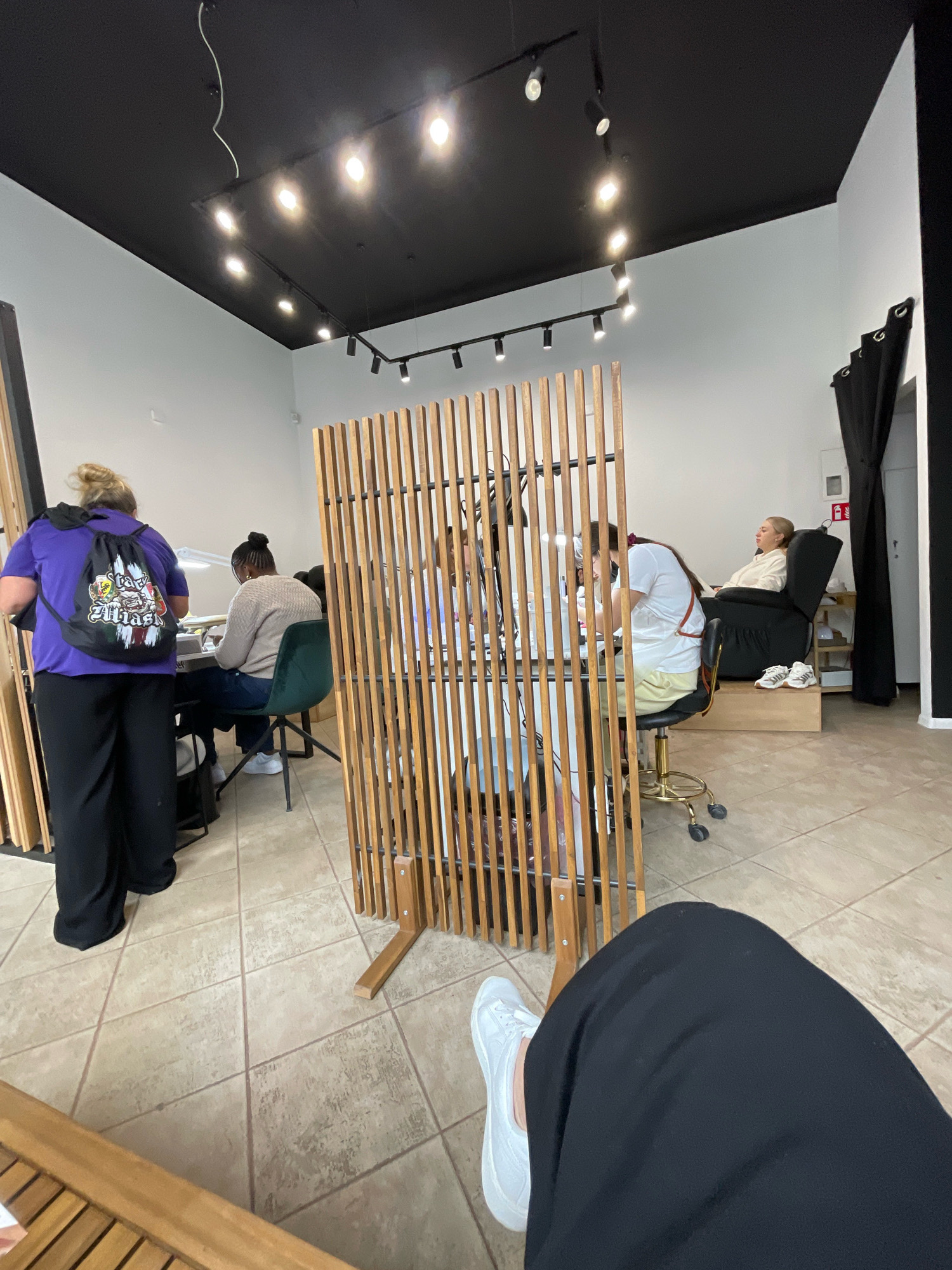 The image depicts an interior scene, likely a nail salon, with several individuals engaged in activities. The room has a black ceiling with modern track lighting. In the foreground, a person wearing black pants and white sneakers is seated, with their legs visible. To the left, another person in a purple shirt and black pants, carrying a backpack, stands near a nail technician's station. A nail technician is seated, working on a client. In the background, a woman reclines in a black chair, possibly receiving a pedicure. The space is decorated with wooden slatted screens, and the floor is tiled. The image provides no information to identify the city as Wrocław, Poland.