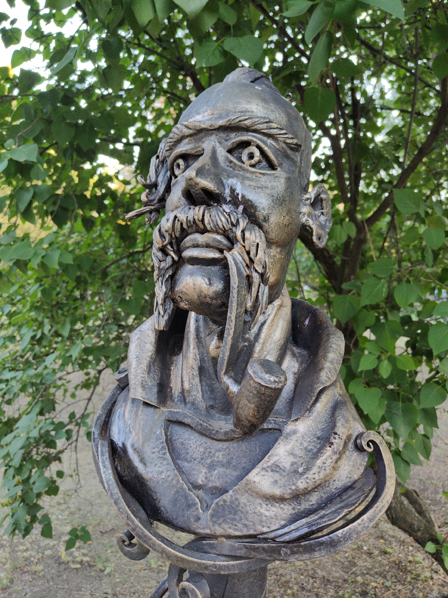 A bronze bust depicts a man with a prominent mustache and beard, smoking a pipe. The sculpture is positioned outdoors against a backdrop of green foliage and tree branches, suggesting a park or garden setting. The man has a stern expression, with defined facial features and his eyes open. He is wearing a cloak or scarf draped around his shoulders. The bust is mounted on a decorative base, partially visible at the bottom of the frame. The overall impression is that of a historical or artistic monument. This bust is located in the city of Donetsk, Ukraine.