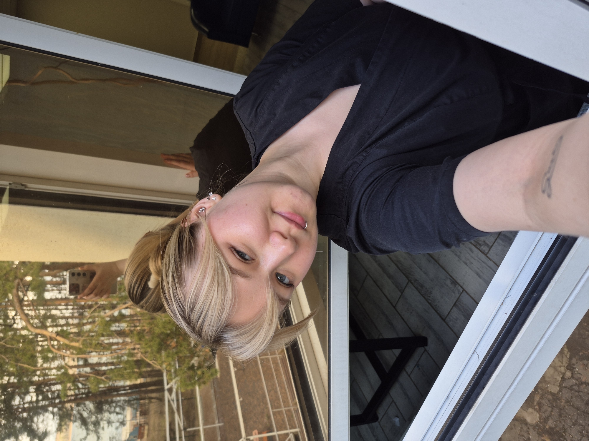 The image displays a young person taking a selfie, with the photo oriented 90 degrees counter-clockwise. The main subject is a fair-skinned individual with blonde and light brown hair, styled with bangs, looking towards the camera with a subtle smirk or playful expression. They have blue eyes, multiple ear piercings (studs and hoops), and a silver ring piercing on their lower lip. They are wearing a black short-sleeved top or shirt, and a dark tattoo is visible on their left forearm.
The person is positioned next to a white window frame, which acts as a barrier between an indoor setting and an outdoor environment. The window glass reflects various elements: to the left of the subject's head, their hand is seen holding a smartphone, which is likely being used to capture the image. The reflection also reveals an outdoor scene featuring numerous trees, possibly conifers, suggesting a natural or suburban area. The warm, golden hue on the reflected trees indicates it is likely daytime, possibly late afternoon.
Within the reflected indoor space, part of a light-colored, possibly tiled floor is visible, along with a dark, angular object that could be a chair leg or other furniture. Outside the window frame on the bottom right, an outdoor ground surface, possibly dirt or gravel, can be seen. The scene captures a casual moment in a building with a view of greenery. No discernible text is visible in the image.
