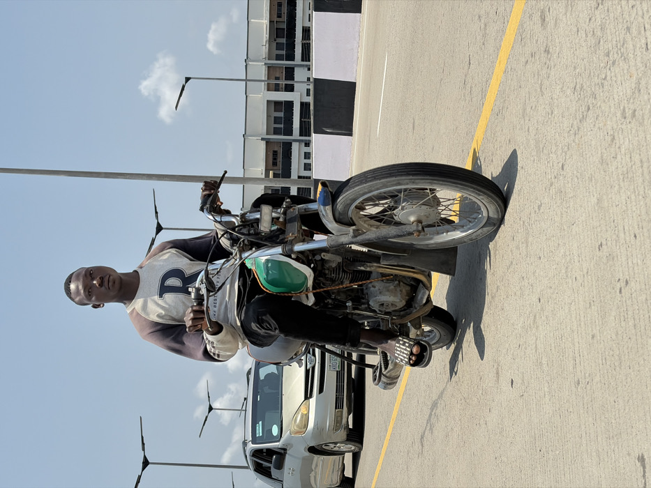 The image displays a young adult male seated on a motorcycle on a paved road in Alaguntan, Nigeria. The male, with dark skin and a short haircut, wears a grey and dark-sleeved raglan shirt featuring a prominent white "R" emblem, black trousers, and black-and-white striped slide sandals. He grips the motorcycle's handlebars and looks directly forward.
The motorcycle is an older model, distinguished by a green and white fuel tank, exposed engine components, spoked wheels, and a visible front fender. A silver Toyota Sienna minivan, bearing the license plate "KRD 659 FK", is positioned behind the motorcycle to the left. The road surface is concrete or asphalt, marked with a solid yellow lane line. A black and white striped concrete barrier runs along the right side of the road. In the background, modern streetlights with flat fixtures are visible on tall poles, alongside multi-story buildings. The sky is clear blue with minimal clouds, indicating bright daylight that casts distinct shadows.