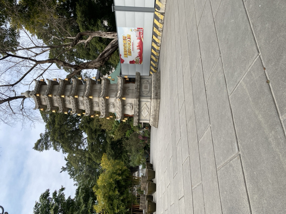 This image captures an outdoor scene featuring a multi-tiered stone pagoda, lush greenery, and a paved walkway. The pagoda, made of light grey stone, stands centrally and features intricate carvings and small golden bells or ornaments hanging from the eaves of each tier. Its base is broad and also carved with decorative patterns.
Surrounding the pagoda are various trees; some have bare branches, suggesting a cooler season, while others are full of vibrant green leaves. In the background, to the right of the pagoda, there is a temporary white wall or barrier with a yellow and black striped base. A large red banner is affixed to this wall. The banner displays Chinese characters, prominently "喜迎二十大" (Xǐ yíng èrshí dà), which translates to "Happily Welcome the 20th National Congress," and "奋进新征程" (Fènjìn xīn zhēngchéng), meaning "Strive forward on a new journey." A stylized red cityscape silhouette is also visible on the banner, along with a blurred series of zeros that appears to be a phone number.
The foreground and right side of the image show a wide paved area composed of rectangular grey tiles. A line of dark, square planters is arranged along the edge of this paved area near the base of the pagoda. The sky overhead is overcast, indicating a cloudy daytime setting. The scene evokes a tranquil, possibly historical, public space or temple ground in Guangzhou City, China.