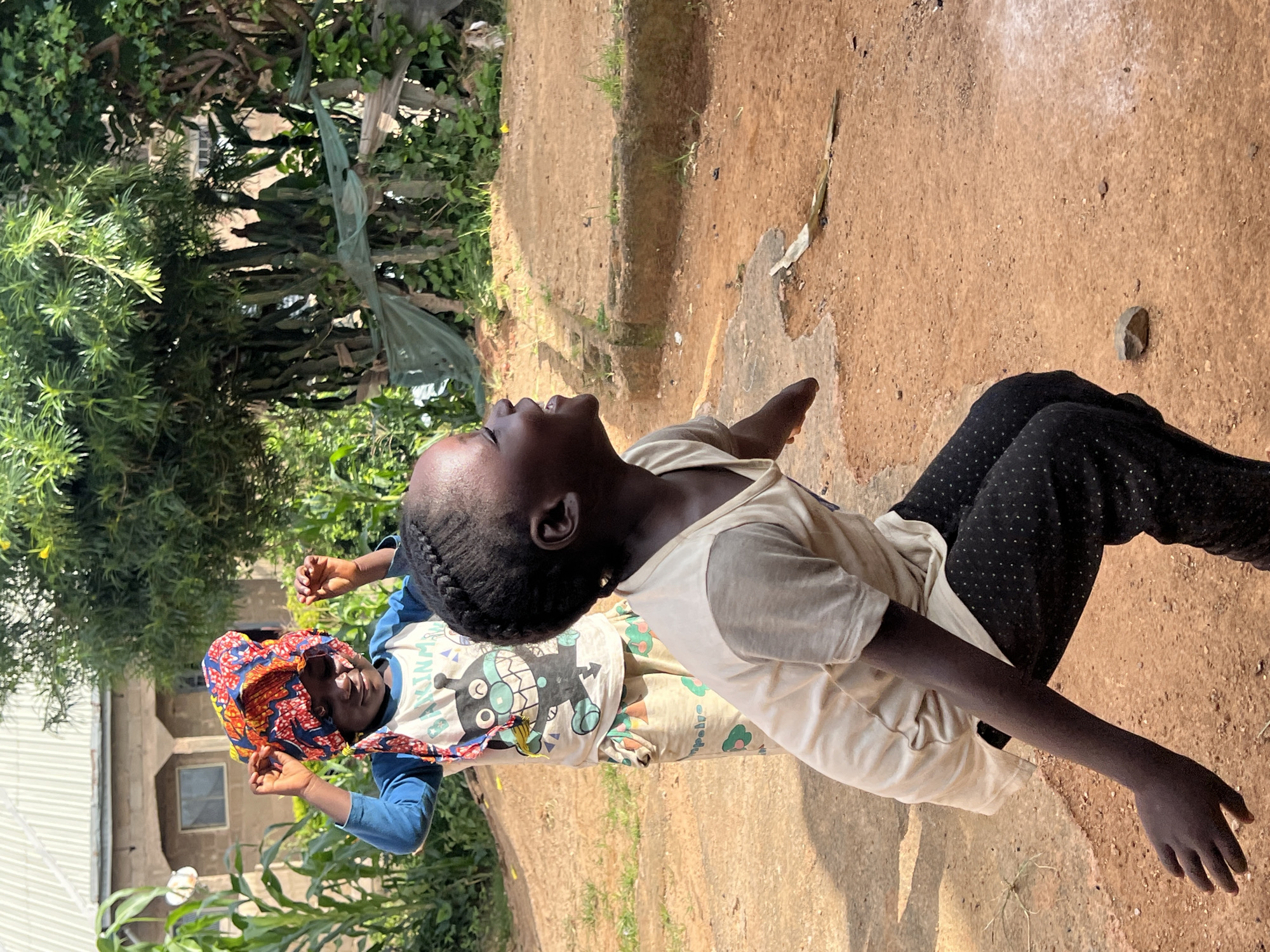 The image shows two young Black girls in an outdoor setting, likely a residential area in Jos, Nigeria. The girl in the foreground, wearing a light-colored t-shirt and black pants with white polka dots, is captured in mid-action, appearing to be jumping or reaching out with her left arm extended. She has a bright, engaged expression, looking upwards and to the right. Her hair is styled in braids. Behind her, another girl is standing, wearing a blue long-sleeved shirt and patterned pants, with a colorful headscarf tied around her head. She is also looking down and to the right, with her hands raised near her head.
The environment is characterized by reddish-brown dirt ground, with some patches of grass and scattered stones. A few concrete structures are visible, suggesting a rudimentary building or foundation. In the background, there is lush green vegetation, including corn stalks and trees, and a building with a corrugated metal roof. The overall impression is a scene of children playing outdoors in a rural or semi-rural area.