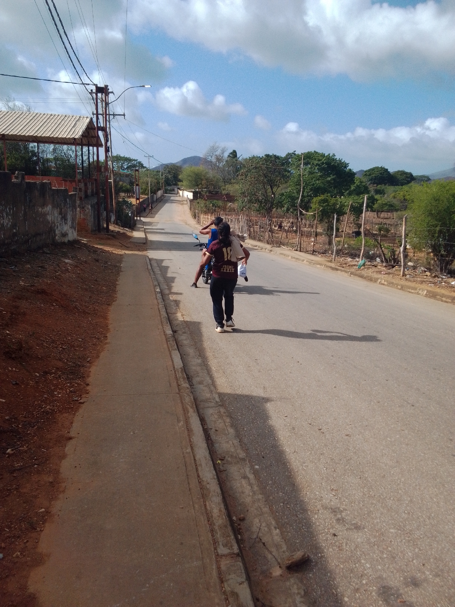 The image depicts a road scene in La Tagua, Venezuela. In the foreground, a woman walks away from the camera on a paved sidewalk adjacent to a dirt embankment. She is wearing dark pants, white sneakers, and a dark t-shirt with "18 PROMO XXXIX" printed on it. She carries a white plastic bag in her left hand. Behind her, on the asphalt road, another person is visible on a motorcycle, with a dark-haired individual seated behind them. The road extends into the distance, lined by a wooden fence and trees. Power lines are strung overhead. A covered structure with a corrugated roof stands on the left side of the image, near a concrete wall. The sky is blue with scattered white clouds. The overall impression is a sunny day in a somewhat rural or semi-urban environment.
