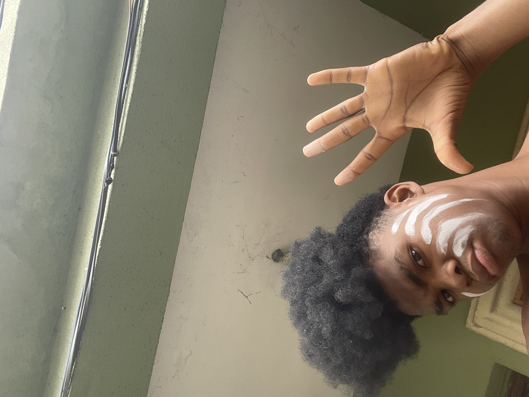 A low-angle selfie of a person with dark skin, afro hair, and white paint stripes on their face. The person's right hand is raised towards the camera with fingers spread. The background shows a light green wall with visible wiring and a lighter-colored ceiling. The location appears to be indoors, possibly in Ado, Nigeria, based on the user's request.
