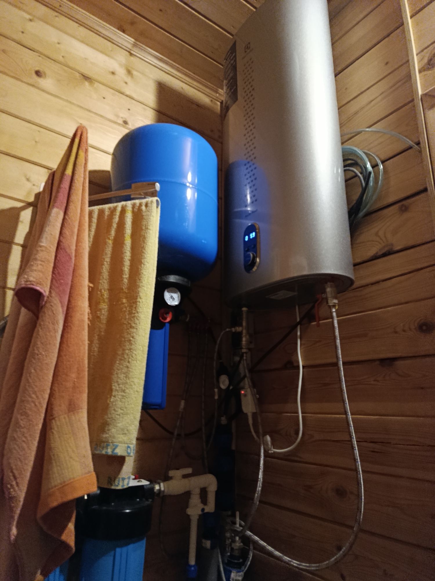 The image depicts a close-up view of a water heating and filtration system mounted on a wooden wall, likely in a utility room or basement in Verezya, Russia. On the left, a blue expansion tank is visible, next to which two towels are hanging on a rack. To the right of the expansion tank is a silver-colored cylindrical water heater with a digital display. Various pipes, gauges, and electrical wires connect the different components of the system. The wooden walls are made of horizontal planks. The scene is dimly lit, suggesting an indoor environment. There are no people or animals visible in the image. The focus is entirely on the mechanical equipment.