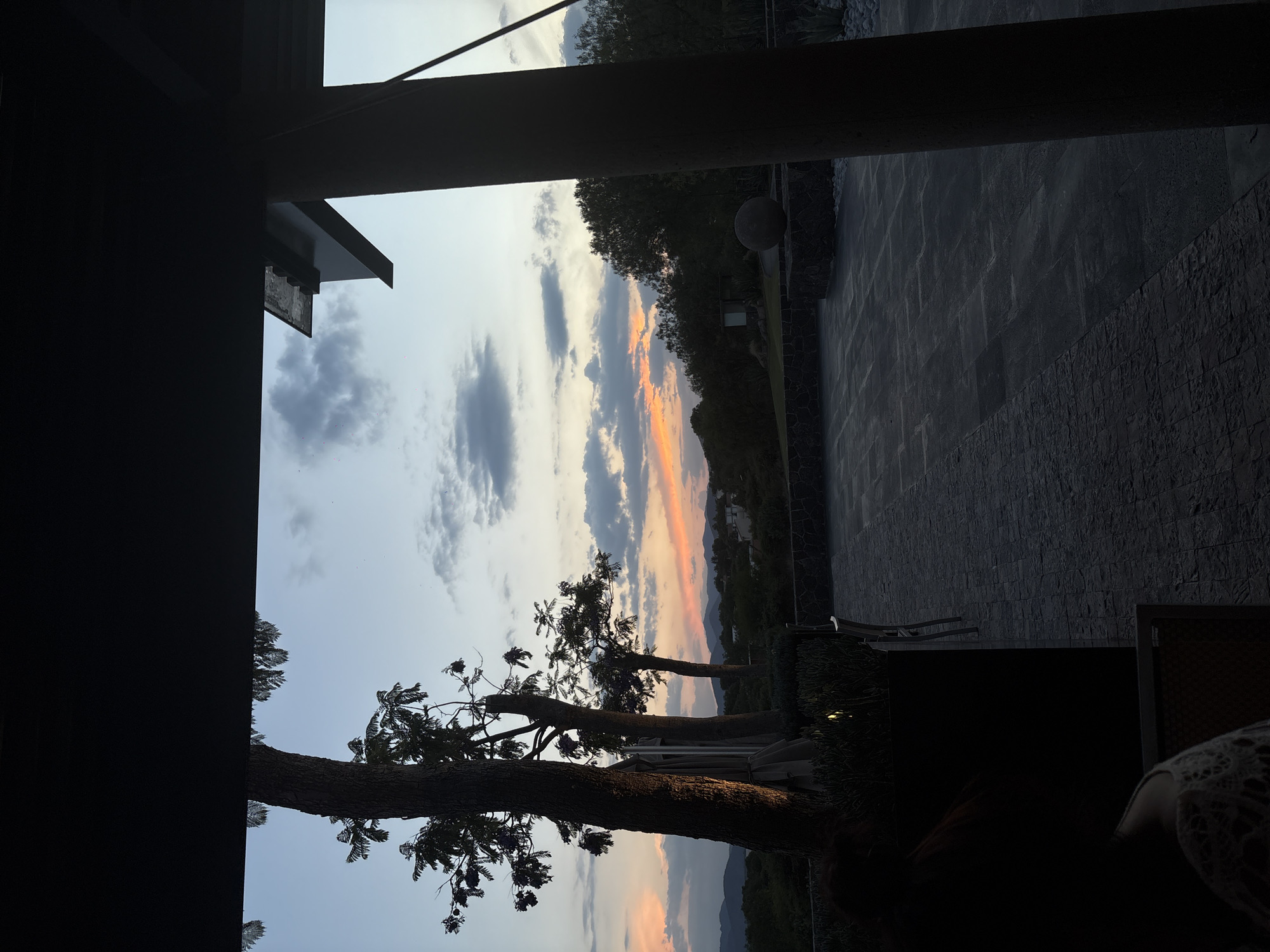 The image presents a view of a sunset in Pueblo Nuevo de Jasso, Mexico. The scene is framed by architectural elements, including dark columns and portions of a building roof, suggesting the vantage point is from within a structure or covered area. The sunset displays a gradient of colors, transitioning from blue skies to orange and pink hues near the horizon. Clouds are scattered across the sky. Three trees are silhouetted against the sky, with their branches extending into the view. A patio area with stone flooring is visible on the right side, with a stone wall extending into the distance. Minimal lighting is present on the property. Darkened areas on the bottom-right corner hint at furniture.