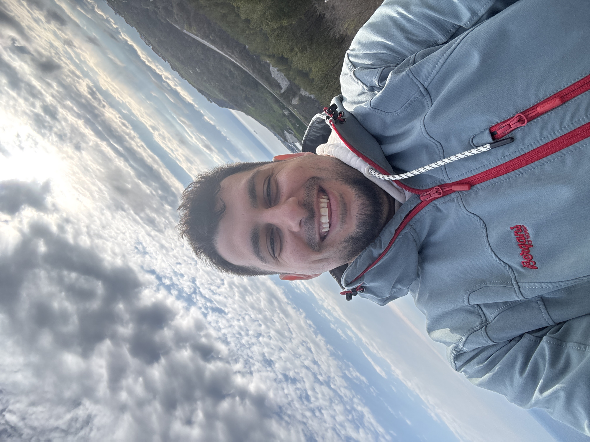 A close-up, rotated selfie depicts a man with short, dark hair and a trimmed beard smiling directly at the camera. He is wearing a light blue Bergans jacket with red zipper accents and a white inner layer. The background reveals a cloudy sky with a glimpse of green vegetation and a body of water, potentially a lake or sea. It is not possible to confirm the specific location as Topola, Bulgaria, with the given information.