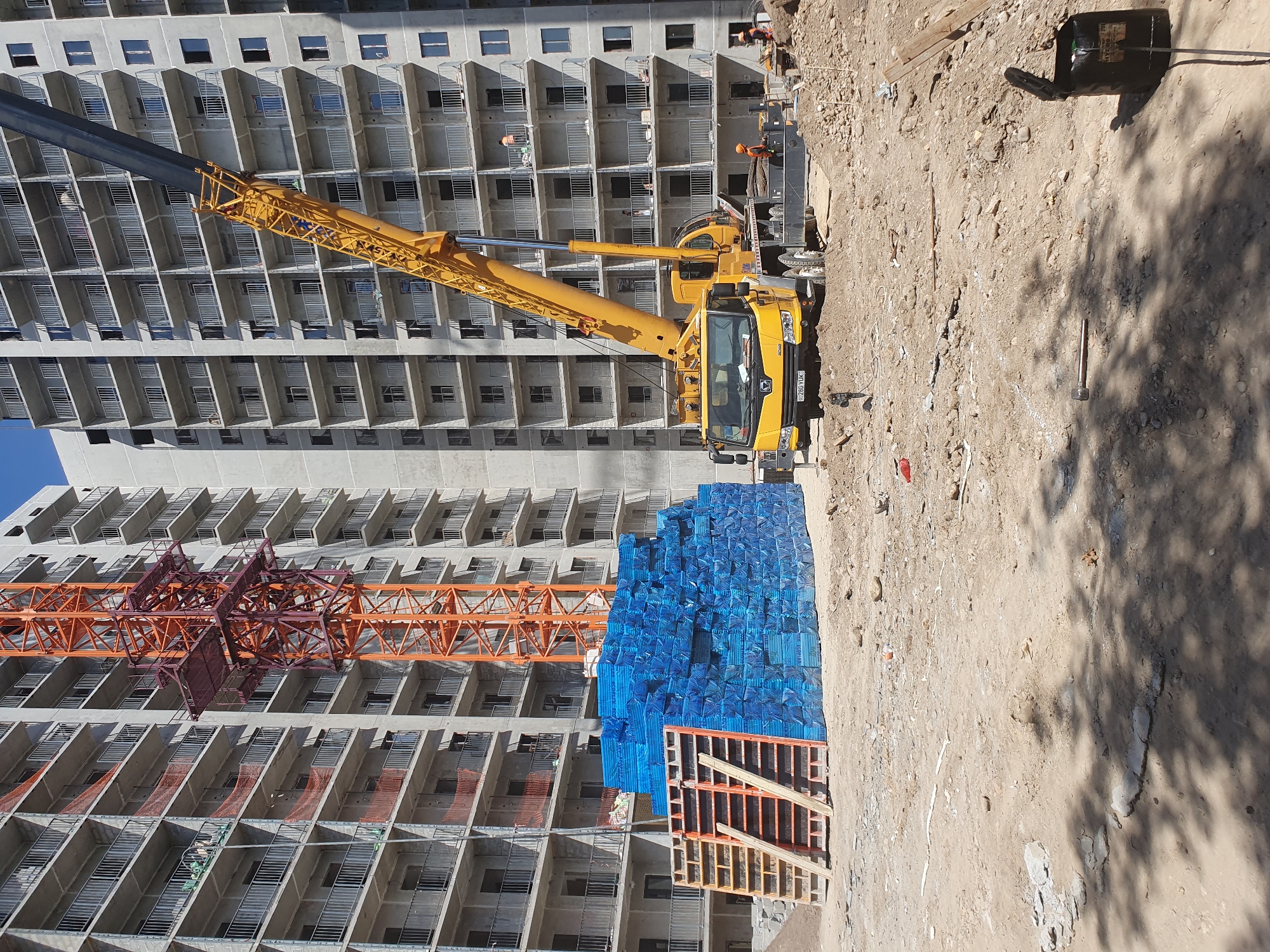 A construction site in Tashkent, Uzbekistan, features a large building under construction with multiple stories and balconies. A tall orange tower crane is partially visible on the left side of the image, and a yellow mobile crane with its boom extended is prominent on the right. In the foreground, stacks of blue-wrapped materials, likely construction supplies, are piled high next to wooden formwork. A yellow truck, possibly a crane vehicle, is parked on the dirt ground. Several construction workers in orange vests and hard hats are present, some working near the mobile crane and others on the balconies of the building. The ground is a mixture of dirt, gravel, and scattered debris. The sky is clear and blue. The scene suggests ongoing construction activities.