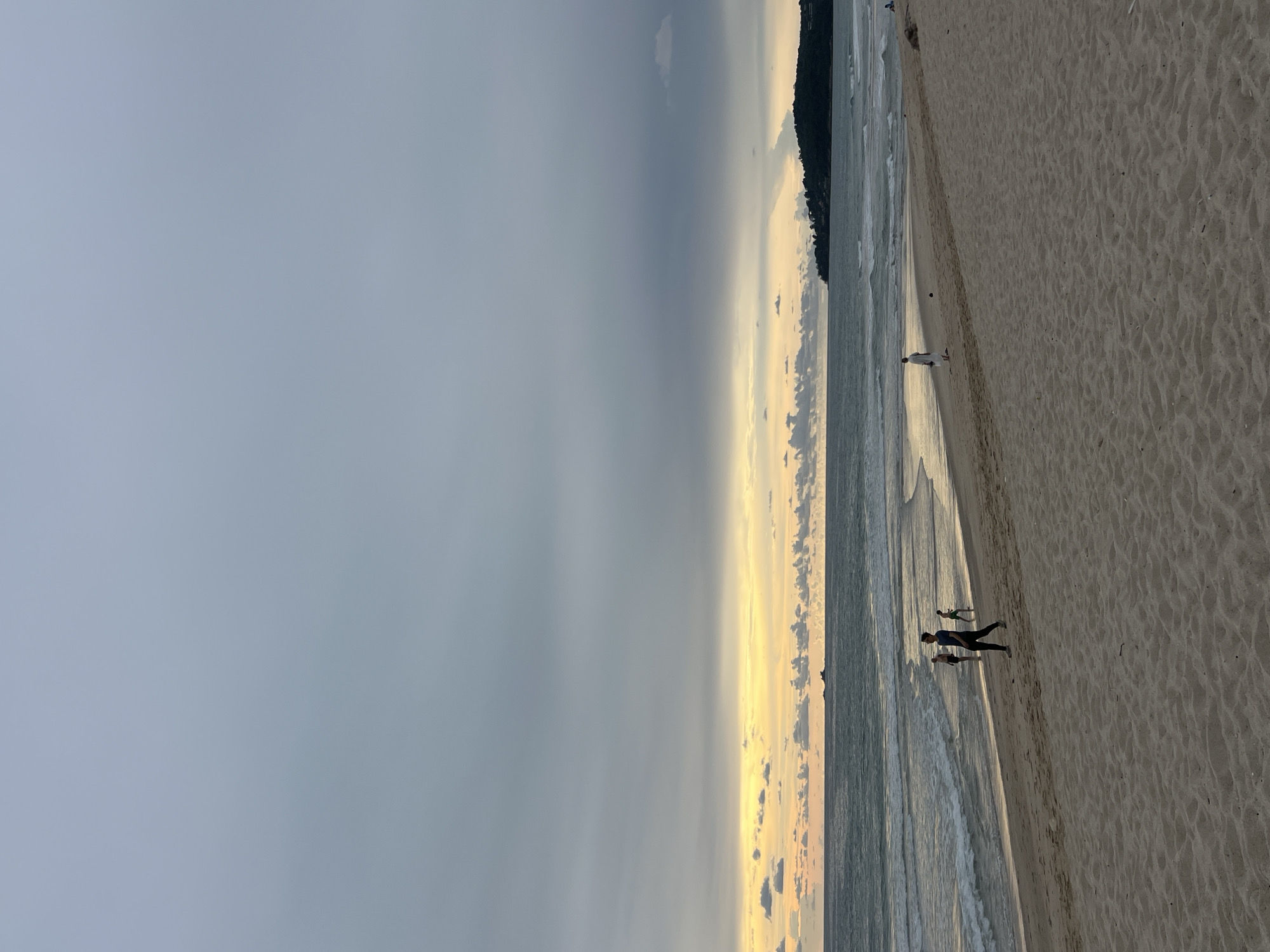 A wide-angle, eye-level shot captures a serene beach scene in Choeng Thale, Thailand, during the late afternoon or early evening. The sky is predominantly a soft, muted grey with streaks of pale yellow and orange near the horizon, indicating the setting sun. A faint, hazy glow emanates from the sunset, casting a warm light over the water and clouds. Dark, wispy clouds are scattered across the upper half of the frame, adding texture to the sky.
The ocean stretches across the middle ground, with gentle waves lapping at the shore. The water appears a muted blue-grey, reflecting the overcast sky. A small, dark island or landmass is visible in the distance on the right side of the frame, topped with lush green foliage.
The foreground is dominated by a sandy beach, appearing pale beige and textured with subtle ripples and indentations. In the mid-ground, several people are scattered across the wet sand near the water's edge and further up the beach. On the left, three individuals are walking or standing near the water, their forms silhouetted against the brighter sky. One person, dressed in dark clothing, is walking further up the beach towards the foreground. To their right, another person, also in dark attire, is walking in the same direction. Further to the left, another figure is partially obscured by waves.
On the right side of the beach, a single individual in white clothing stands near the water, facing the ocean. Another person, dressed in white, is walking on the sand further up the beach. The presence of these individuals suggests a tranquil atmosphere, with people enjoying the last light of day on the beach.