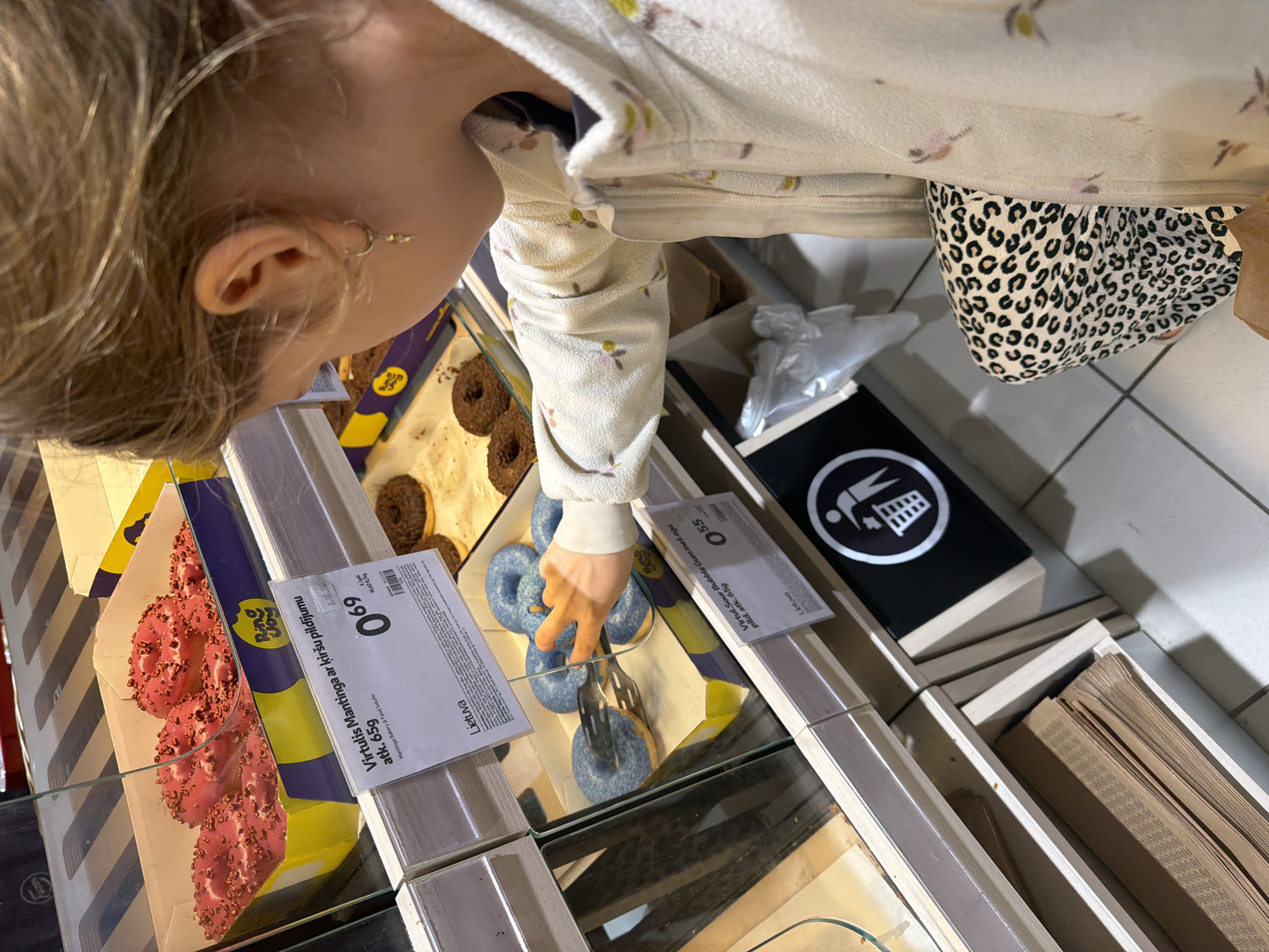 A young child, observed from behind and the right side, is actively selecting a donut in a grocery store located in Krustkalni, Latvia. The child's light-colored hair is pulled back, revealing a small, gold-toned hoop earring with a dangling charm on the right ear. They are wearing a cream-colored fleece jacket adorned with small pink and yellow floral patterns, and leopard-print pants featuring black spots on a light background.
The child's right hand, encased in the jacket sleeve, holds a pair of silver metal tongs, which are being used to grasp a blue-frosted donut from a display case. This donut is situated within a yellow and purple cardboard box containing other blue-frosted donuts. A price label in front of this box reads "Virtulis Sour Bubble Gum mežogu pild. atk. 65g" (Sour Bubble Gum Donut with berry filling approx. 65g) and displays a price of "0.55," alongside the country of origin "Lietuva" (Lithuania).
Above this, another glass-covered display section contains a yellow and purple cardboard box filled with multiple pink-frosted donuts topped with dark sprinkles. The corresponding price label states "Virtulis Mantinga ar ķiršu pildījumu atk. 65g" (Mantingas Donut with cherry filling approx. 65g) and shows a price of "0.69," also indicating "Lietuva" and the brand "Ring Joy." To the right of the blue donuts, a clear glass tray holds several chocolate-frosted donuts.
Below the display cases, a stack of flat brown paper bags is neatly stored in an open wooden compartment, presumably for packaging baked goods. Adjacent to this, a black container with a white circular icon depicting a person discarding trash into a bin is visible. The floor consists of light-colored tiles, and the setting is illuminated by artificial indoor lighting.