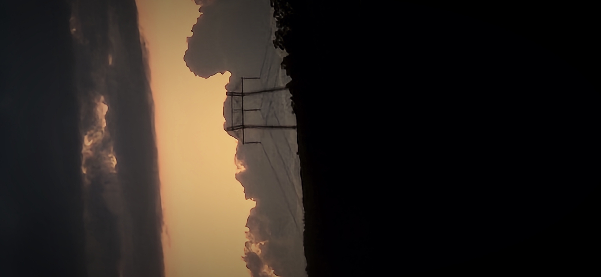 The image displays a landscape scene at sunset in Gutsal, Bulgaria. The upper third of the frame consists of a dark, heavy cloud layer, predominantly deep grey and black, with subtle lighter, reddish-brown highlights along its lower edges. Below this, the sky transitions into a vivid band of orange and yellow, indicating the setting sun.
A prominent electrical transmission tower, rendered in dark silhouette, stands centrally within the mid-ground, extending from the dark foreground into the vibrant orange sky. Its structure is identifiable as a large metal lattice pylon with a broad base and a multi-tiered cross-arm section at its apex. Faint, lighter lines, consistent with overhead power cables, are visible extending from the pylon's cross-arms, partially obscured by distant clouds.
Behind the pylon, a mass of lighter grey clouds or distant, elevated terrain forms a wavy silhouette against the bright sky. To the far right, a cluster of dark, silhouetted trees or dense foliage is discernible. The entire lower half of the image is an undifferentiated expanse of deep black, representing the foreground landscape in extreme shadow, with no specific details visible. No individuals or discernible actions are present in the scene.