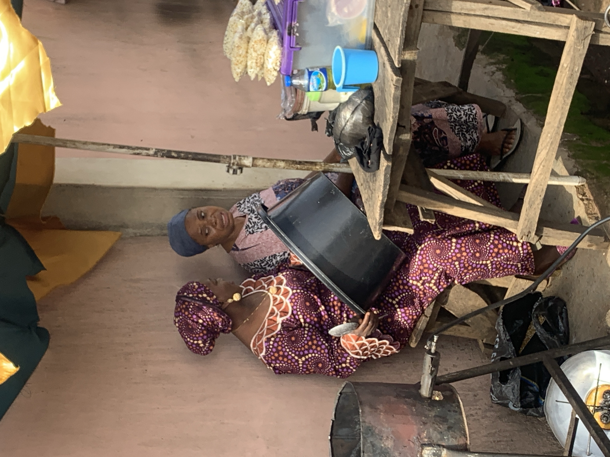 The image depicts two women in what appears to be an outdoor or semi-covered market setting in Jos, Nigeria. The woman in the foreground is wearing a patterned purple and orange headwrap and a matching dress with intricate white and orange detailing around the neckline. She is looking downwards, holding a large, dark rectangular object, possibly a tray or a lid, in her hands. She is also wearing a gold necklace. To her left, further back, another woman is visible, wearing a blue headwrap and a patterned pink and white top. She is looking towards the camera with a slight smile.
The scene is cluttered with various objects, suggesting a market stall or storage area. There are wooden planks forming a structure, possibly a table or shelter. On the wooden structure, there are several items, including a blue cup, a bottle of water, and bags of what look like snacks or food items. There is also a black garbage bag and a metal drum or cooking pot visible. The ground is a dusty, light-colored surface.
The lighting suggests it is daytime. The overall atmosphere appears to be one of everyday commerce or domestic activity. The women's expressions are neutral to pleasant, indicating no distress. The presence of the food items and the cooking vessel hints at a food-related business or preparation. The orientation of the image is unusual, with the subjects appearing to be lying down.