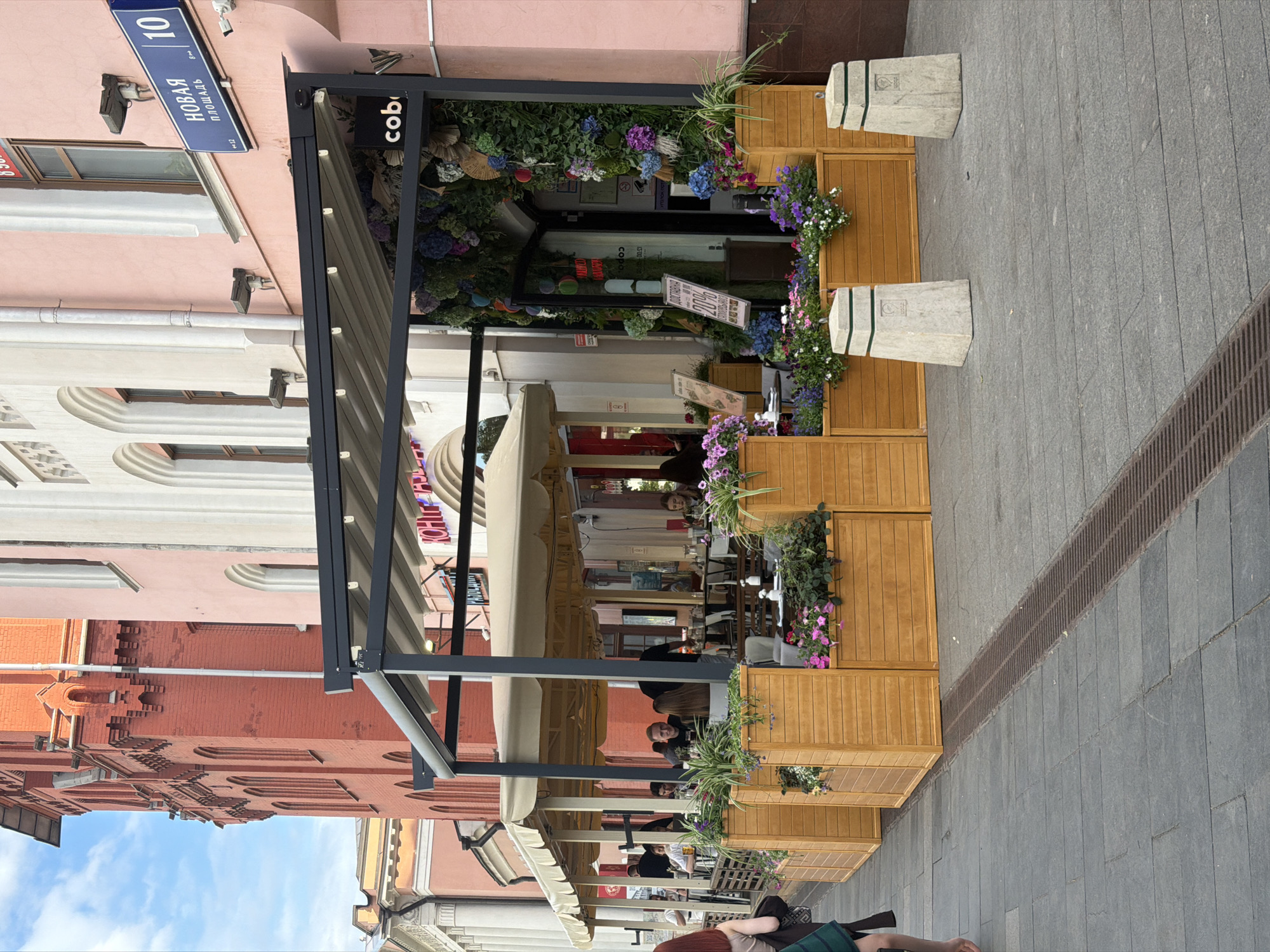 The image depicts an outdoor cafe located on a street in Moscow, Russia, indicated by the "Новая площадь" (Novaya Square) street sign with the number 10. The cafe is partially enclosed with wooden panels adorned with flowers. People are seated at tables under a retractable awning. The architecture surrounding the cafe includes buildings with pink and red facades. The street is paved with grey stone tiles and has a drainage grate running along its edge.