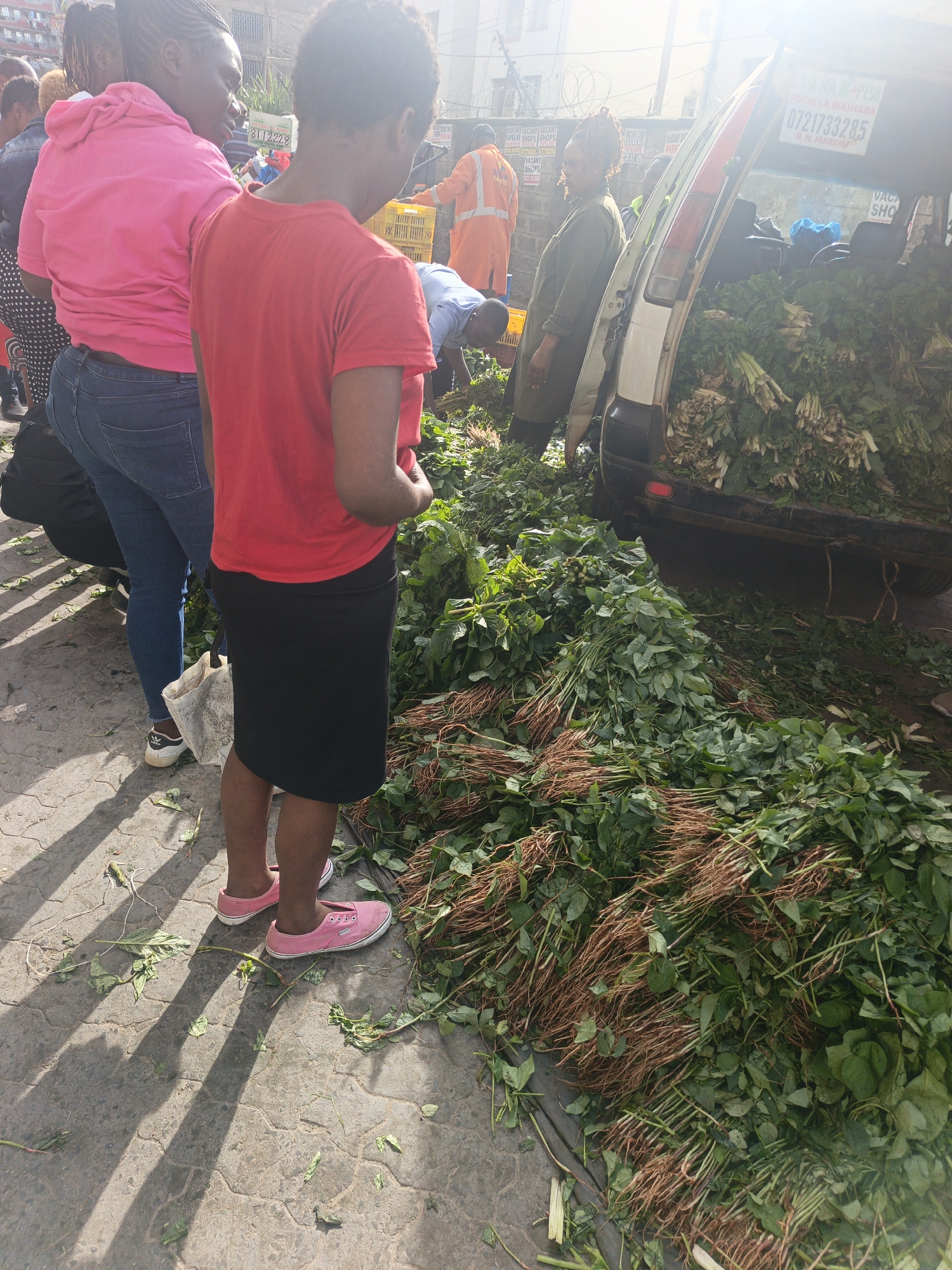 The image shows a roadside vegetable vending scene in what appears to be Nairobi, Kenya, given the visible Swahili signage and Kenyan phone number. A white van is parked with its back doors open, and the interior is filled with leafy green vegetables. More vegetables are piled on the ground around the van. Several people are present. A woman in a pink hoodie and jeans and a woman in a red t-shirt and black skirt are visible in the foreground. An individual wearing an orange work suit is working near the back of the van, presumably unloading or organizing the vegetables. Another woman is standing by the van. The scene is taking place on a paved surface, possibly a street or sidewalk, and sunlight is visible, indicating it is daytime.