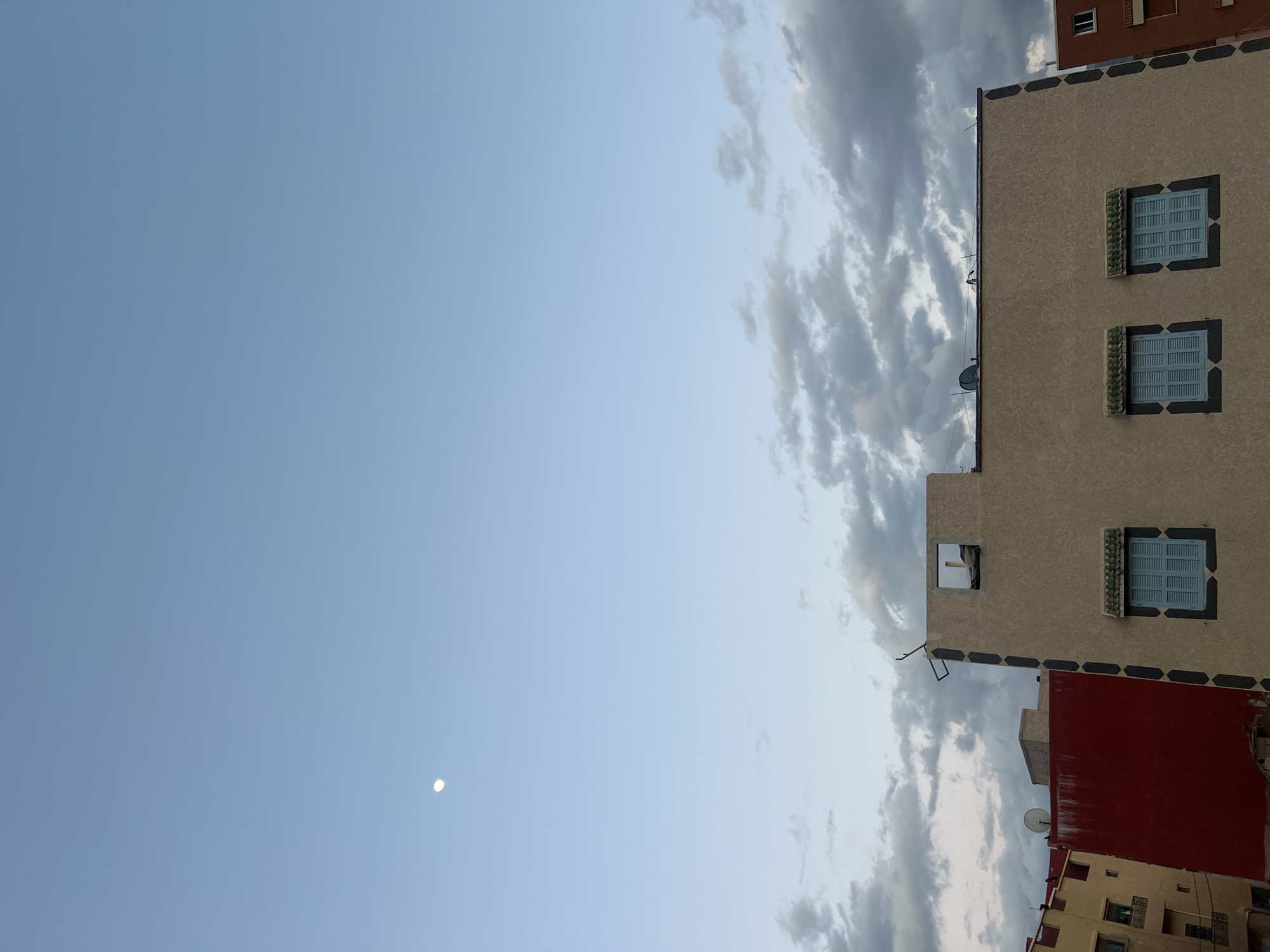 The image presents a view of the sky and the upper portions of buildings in Ouaouizert, Morocco. The sky, predominantly light blue, contains a small, pale orb in the lower left, likely the moon. A band of striated clouds stretches along the right side of the frame. Adjacent to the clouds is a building with a beige facade, decorated with three windows fitted with blue shutters and a distinct patterned trim. Above this beige building, a portion of an orange/red building is also visible. Further below, the top portion of another building, lighter in color is observable. A satellite dish is affixed to one of the roofs. The photograph's orientation appears to be rotated, presenting the scene in a vertical format where the sky dominates the left side.