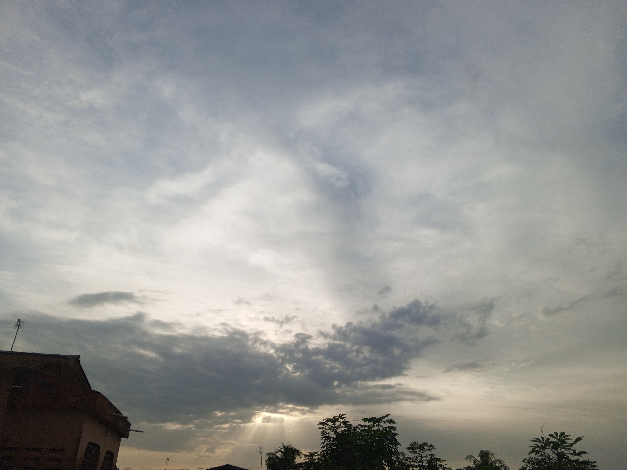 The image displays an outdoor scene dominated by an overcast sky, with elements of a building and vegetation in the foreground. The sky is filled with varying shades of grey clouds, ranging from light, wispy formations to denser, darker clusters. A prominent feature is the dramatic display of sun rays (crepuscular rays) breaking through a gap in the clouds near the bottom center of the frame, indicating the sun is either rising or setting, or obscured by clouds.
In the lower left corner, a portion of a building is visible, featuring a reddish-brown roof, possibly tiled, on which a thin antenna with horizontal elements is mounted. The walls of the building are light-colored, and some architectural details can be seen. Below the sky, along the bottom edge of the image, dark green foliage from trees and bushes is visible, suggesting a natural environment bordering the built structure. These plants partially obscure the lower cloud cover and the sun's rays.
The scene captures a moment of dramatic lighting under a cloudy sky. There are no visible people or discernible text. The combination of the building, antenna, and tropical-looking vegetation is consistent with an urban or semi-urban setting like Badagry, Nigeria.