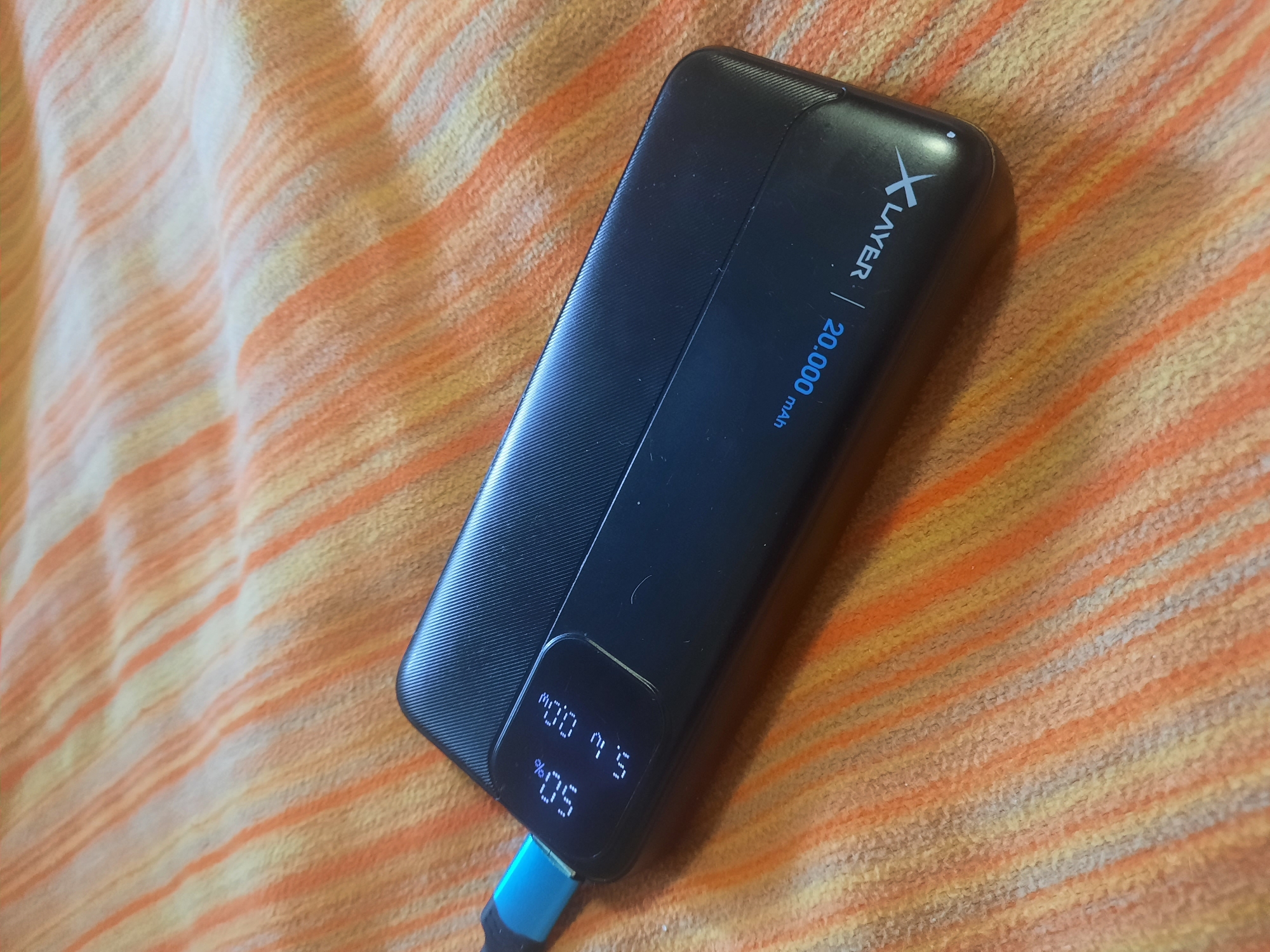 An overhead, close-up shot shows a black power bank with a digital display on a textured, orange and cream striped blanket. The power bank is angled diagonally from the bottom left to the top right of the frame. A blue USB cable is plugged into the left side of the power bank. The digital display shows "50%" and "5.0V / 2.00A". Text on the power bank reads "X LAYER | 20,000 mAh".