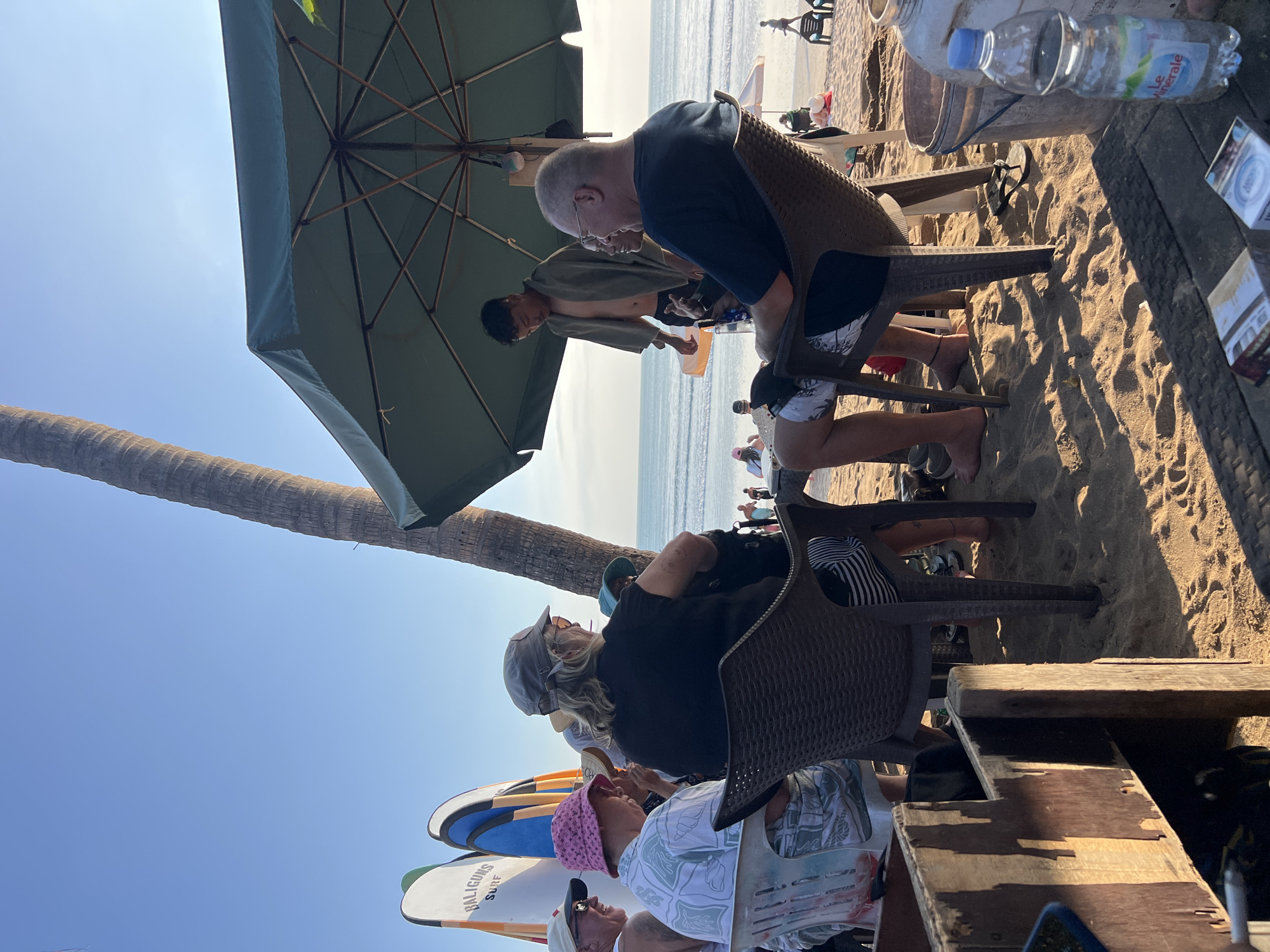 On a sandy beach in Kuta, Indonesia, a group of individuals are gathered near surfboards. In the foreground, four people are seated at a table under a large green umbrella. Two men and two women are present. One man, wearing glasses and a dark blue t-shirt, is facing the camera. Next to him, a woman with blonde hair and sunglasses is looking away from the camera. A woman in a pink hat is to her left, also looking away. Another man, partially obscured, is seated to the far left. A younger man, wearing a towel around his shoulders, stands behind the seated group, appearing to interact with them. In the background, the ocean is visible with gentle waves, and other beachgoers can be seen relaxing on lounge chairs. Surfboards stand upright on the sand to the left of the group. The scene is bathed in sunlight, indicating it is daytime.