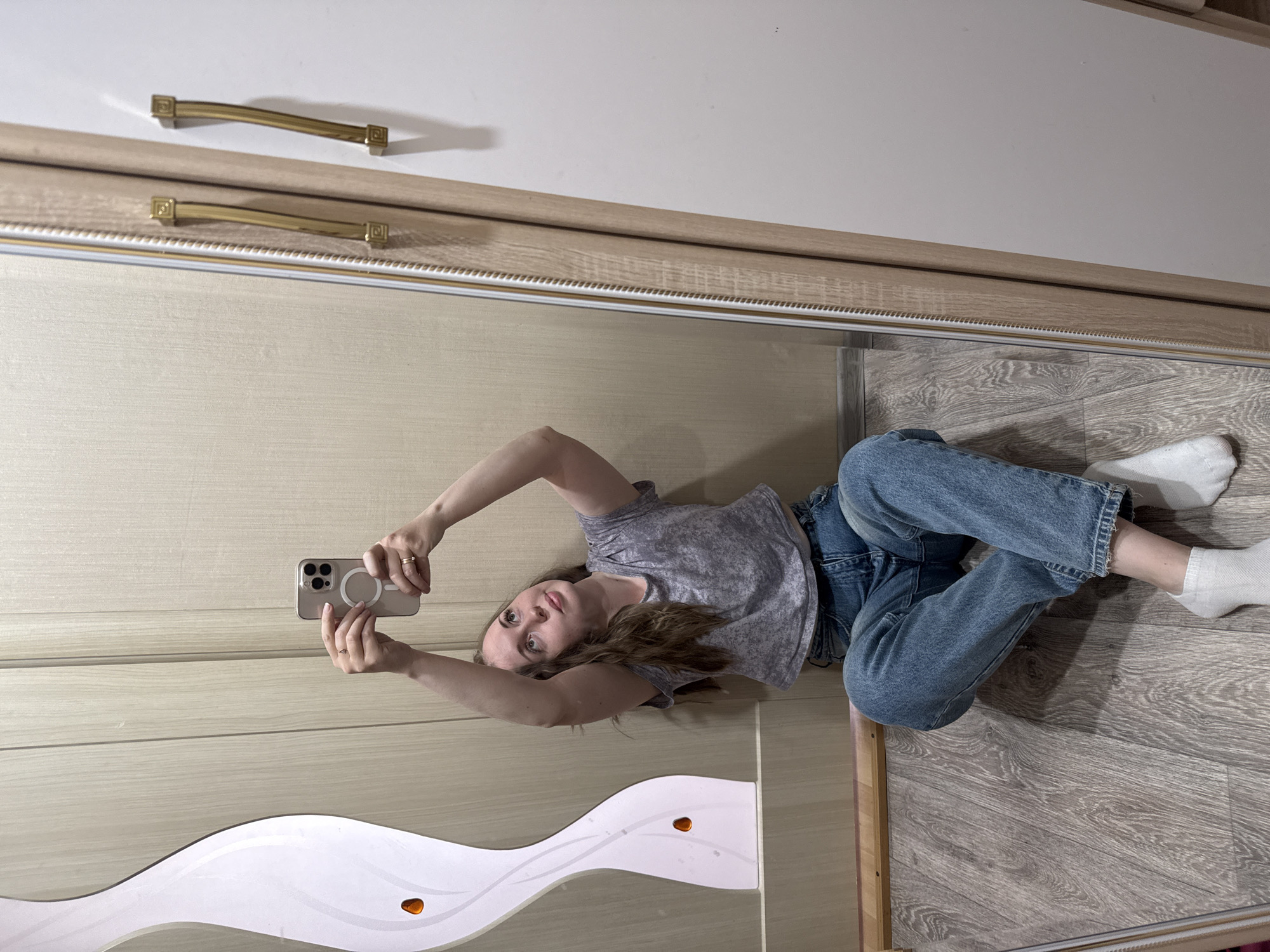 A woman is reflected in a full-length mirror, angled to appear as though she is lying on her side on a wood-patterned floor. She is wearing a gray t-shirt, blue jeans, and white socks. She is holding a smartphone in her hands, presumably taking the picture. The mirror is framed with a light wood trim and attached to a wardrobe with white and wood-paneled doors. The wardrobe features two gold-colored handles. The location is Unknown, Unknown.