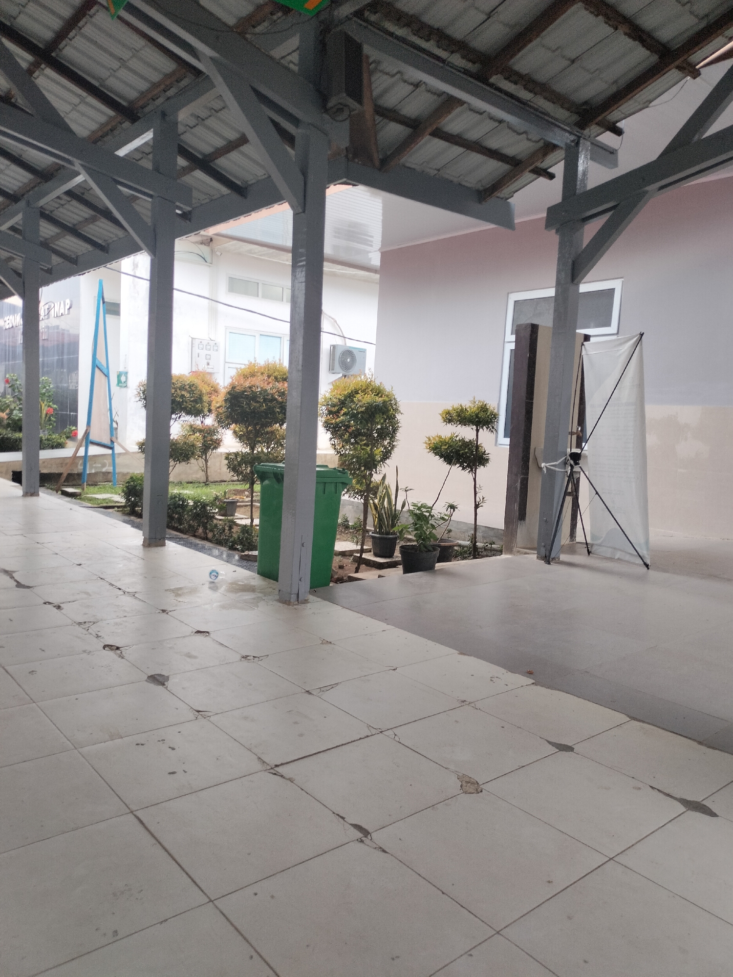 The image depicts an outdoor walkway with a covered roof supported by grey pillars. The walkway is paved with cracked and worn white tiles. To the left, there are several potted plants and trees, as well as a green trash can. Further back, a building with white walls and windows is visible, along with an air conditioning unit mounted on the exterior. On the right side of the walkway, there is a white banner stand on a tripod. The scene appears to be in a public area, possibly a school or hospital, given the signage visible in the background that includes "KEDIK AP NAP". The location is identified as Langsa, Indonesia.