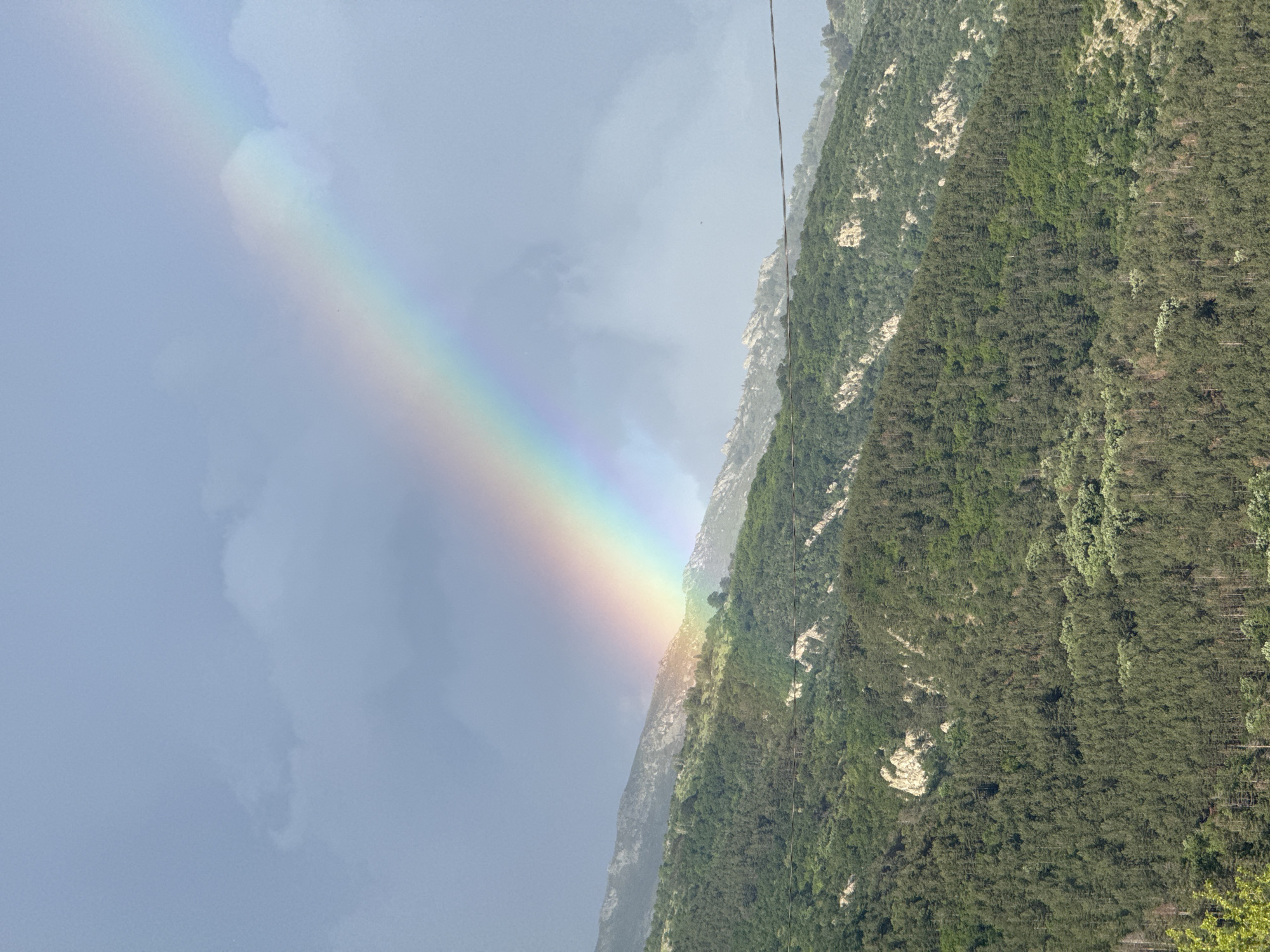 The image depicts a vibrant rainbow ending on a mountainside covered in lush green vegetation. The sky is overcast with varying shades of gray. A cable or wire is visible descending from the top of the frame along the mountainside.