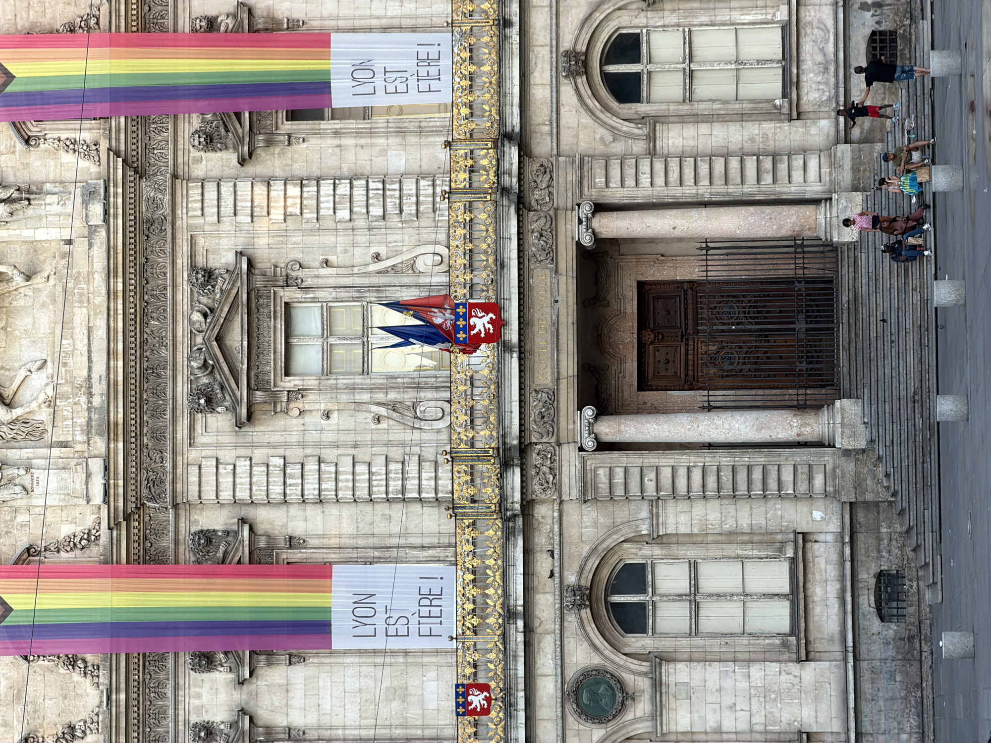 The image captures the facade of Lyon's Hôtel de Ville, also known as the City Hall. Two large rainbow banners hang prominently on either side of the building's central section. Below the banners, a smaller banner reads "LYON EST FIÈRE!". A golden ornate railing runs across the balcony. Two flags, one bearing the coat of arms of Lyon, are affixed to the railing. In front of the building, a set of wide stone steps leads up to the main entrance. Several individuals are visible on the steps and in the plaza in front of the building. One person stands on a step, gesturing with their arms, while others are seated on the steps. The architecture is grand and ornate, typical of classical French design, with carved details and large windows. The setting appears to be daytime, with moderate lighting.