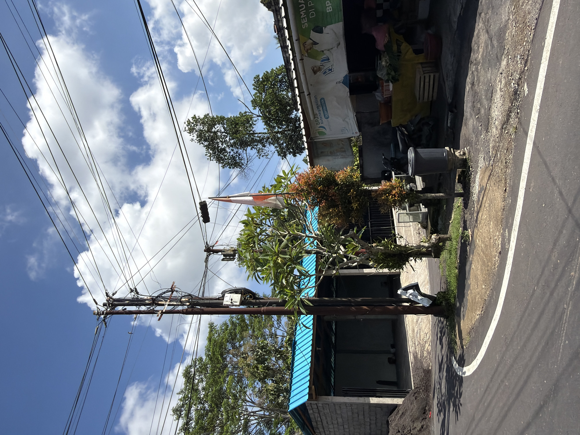 The image depicts a street scene in Tampaksiring, Indonesia, under a clear blue sky with scattered white clouds. A prominent utility pole with numerous wires crisscrosses the frame. To the left, a building with a bright blue corrugated metal roof and concrete block walls is visible. Adjacent to it is a pile of dark soil. In the center, a small tree with green and reddish foliage stands near a tall utility pole. Further to the right, another building is partially visible, featuring a banner with text and images, possibly a storefront. A motorcycle is parked in front of this building. A dark gray trash bin is situated on the sidewalk. The road in the foreground is paved, with a white line marking its edge. The environment appears to be a rural or semi-urban area, characterized by the presence of utility infrastructure, buildings, and vegetation.
