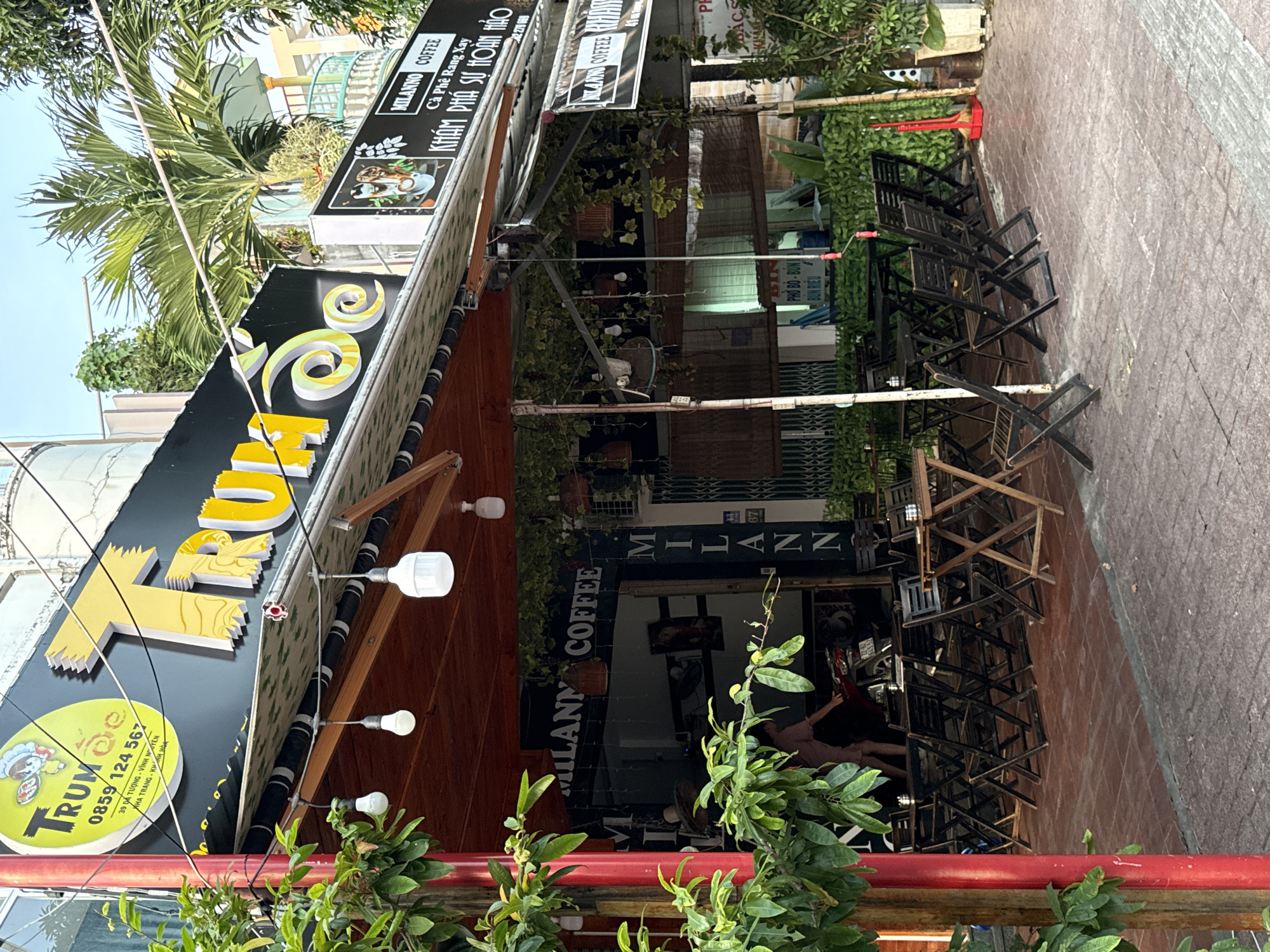 The image shows a street-level view of "Trùm Cà phê" restaurant and coffee shop located at 39 Dã Tượng, Vĩnh Nguyên, Nha Trang, Vietnam. The shop features a prominent yellow sign with the restaurant's name and a phone number, "0859 124 567". A large yellow sign with the word "TRUM" is also visible, with decorative white and yellow lettering. An awning covers the outdoor seating area, which is furnished with dark wooden tables and chairs. A person is partially visible inside the shop, near a motorcycle. The scene is set outdoors with trees and foliage around the establishment, indicating a tropical climate. The sky is visible above.