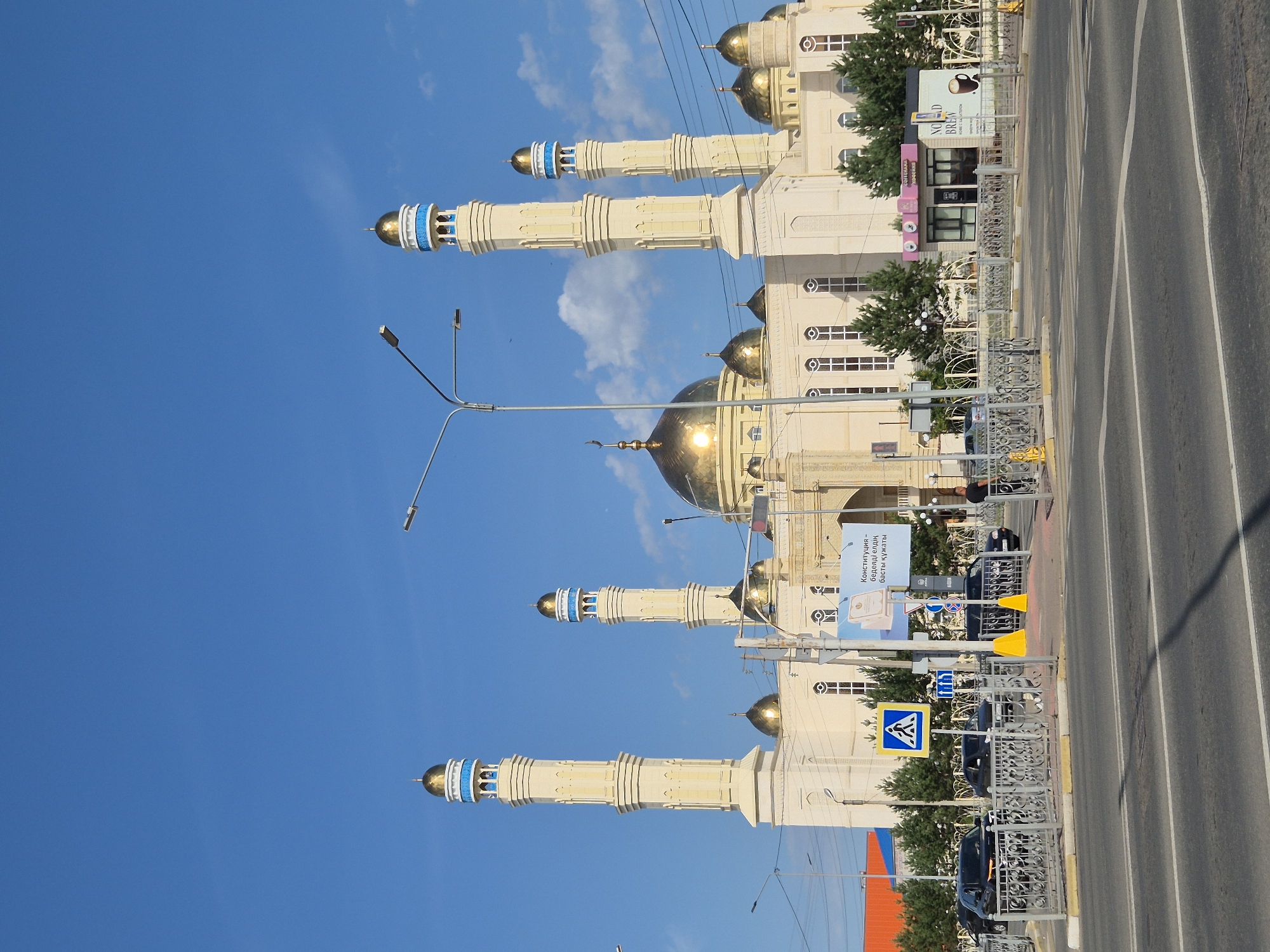 The image depicts a large, ornate mosque with a prominent golden dome and multiple minarets against a bright blue sky. The mosque's exterior is cream-colored with intricate architectural details. In the foreground, there is a street with white lane markings, and a crosswalk. Several black cars are parked or driving near the mosque, and there are pedestrian crossing signs and traffic signals. A fence separates the street from the mosque's grounds, where a few people are visible, including a man in a black t-shirt standing near a fence. A small building with the word "COFFEE" is also visible to the right of the mosque. The mosque in the image is the "Nur Astana" Mosque, located in Kokshetau, Kazakhstan.