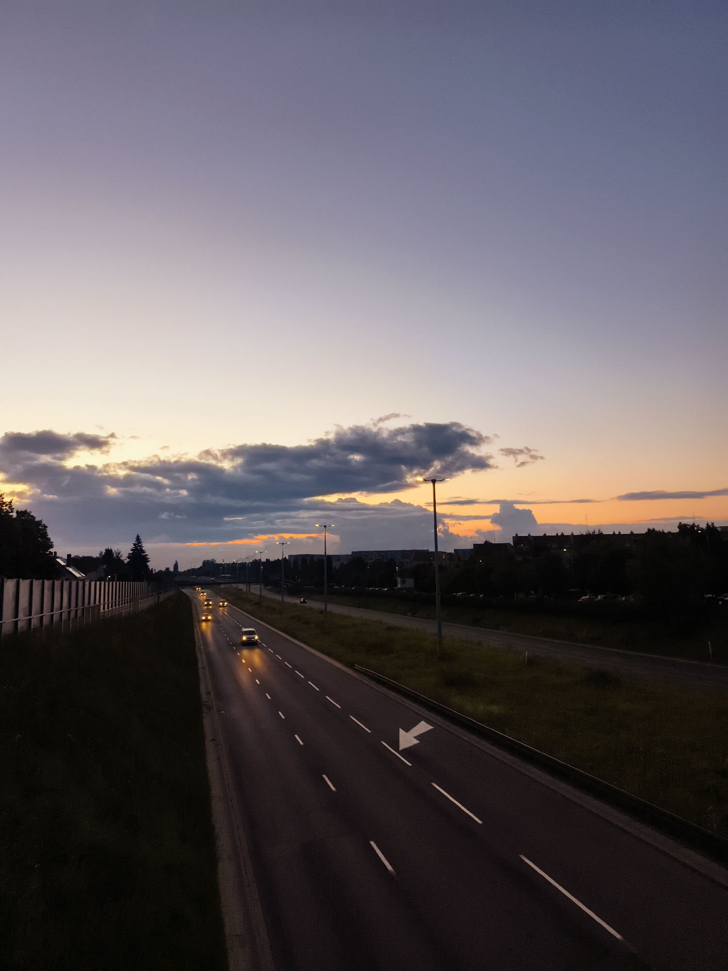 This is a photograph taken at dusk, looking down a multi-lane highway that runs through Gdańsk, Poland. The sky is a gradient of purple, pink, and orange, with scattered dark clouds. The highway is wet, reflecting the headlights of several cars driving in the distance. The lanes are marked with white lines, and a white arrow indicates a lane change. On the left side of the highway, there is a grassy embankment topped with a sound barrier fence. On the right side, trees and buildings are visible in the dim light. Streetlights are positioned along the highway.