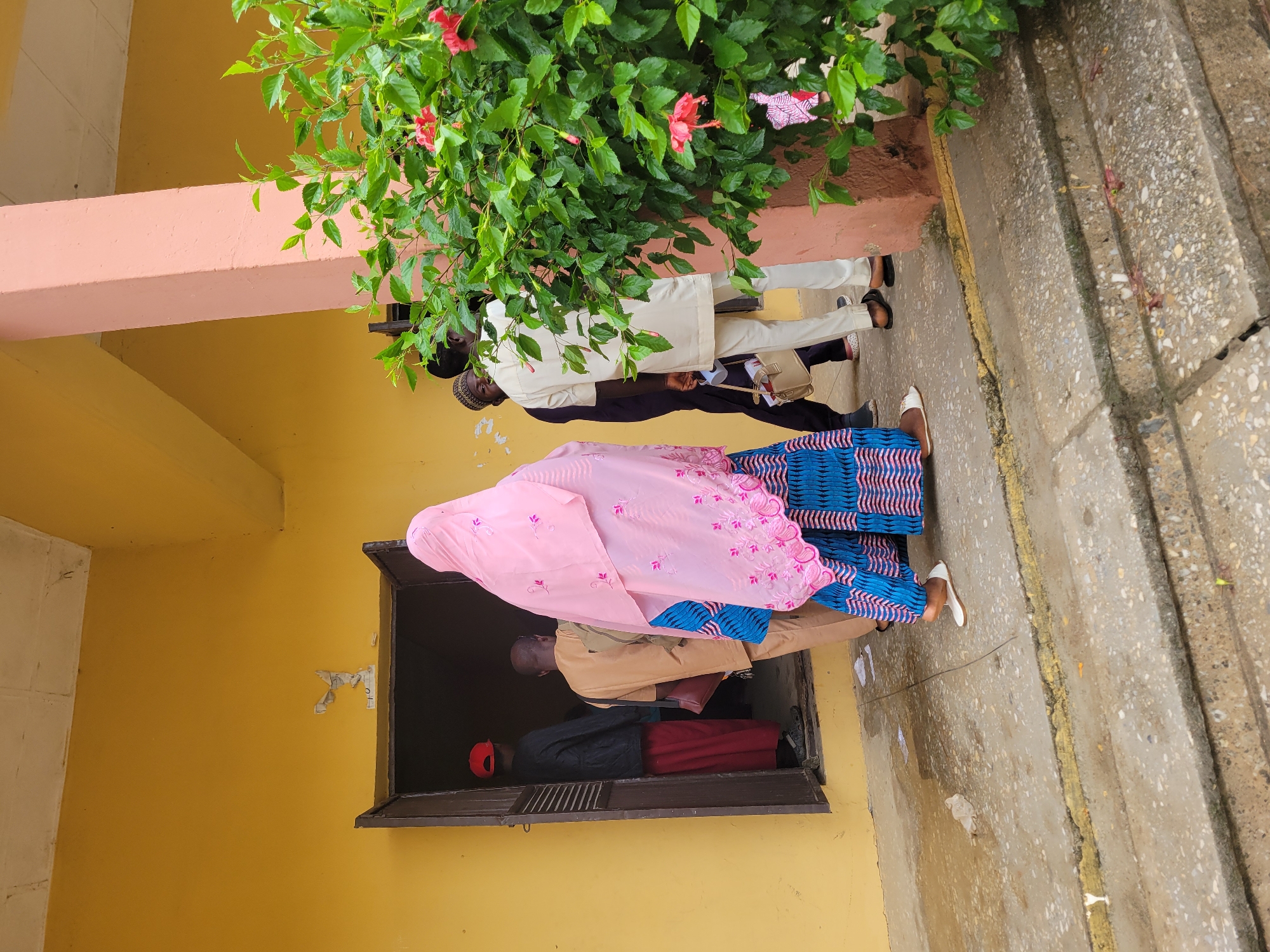 The image captures an outdoor scene in Keffi, Nigeria, depicting the entrance to a yellow-walled building with several individuals. The foreground features a weathered concrete pathway and two steps, with a faded yellow line marking the edge of the upper landing. Small pieces of white paper and plastic debris are scattered on the ground.
A dark, open doorway in the yellow wall is the central focus, with people either entering or queuing to enter. Visible through the doorway, a person wearing a red cap, dark jacket, and red trousers is moving further inside, with their back to the viewer. Directly in front of the doorway, a bald individual in light brown traditional attire with a dark shoulder bag is also facing away, appearing to enter.
In the immediate foreground, a person, likely female, is prominently featured from behind. They wear a light pink head covering adorned with embroidered patterns and a blue and pink patterned dress. White flat shoes are visible on their feet as they move towards the doorway.
To the right, partially obscured by a dense green bush with several red hibiscus flowers, stands another person in light cream/beige traditional tunic and trousers. This individual holds a light beige bag and possibly a white cup or container in their left hand, and wears a dark cap. Their back is to the viewer, facing towards the doorway. Behind them, a pinkish-peach colored pillar rises from the ground, framing the right side of the scene, with another individual partially visible in dark clothing. The building's ceiling, visible at the top, appears to be a simple, light-colored structure. The scene is illuminated by natural daylight.