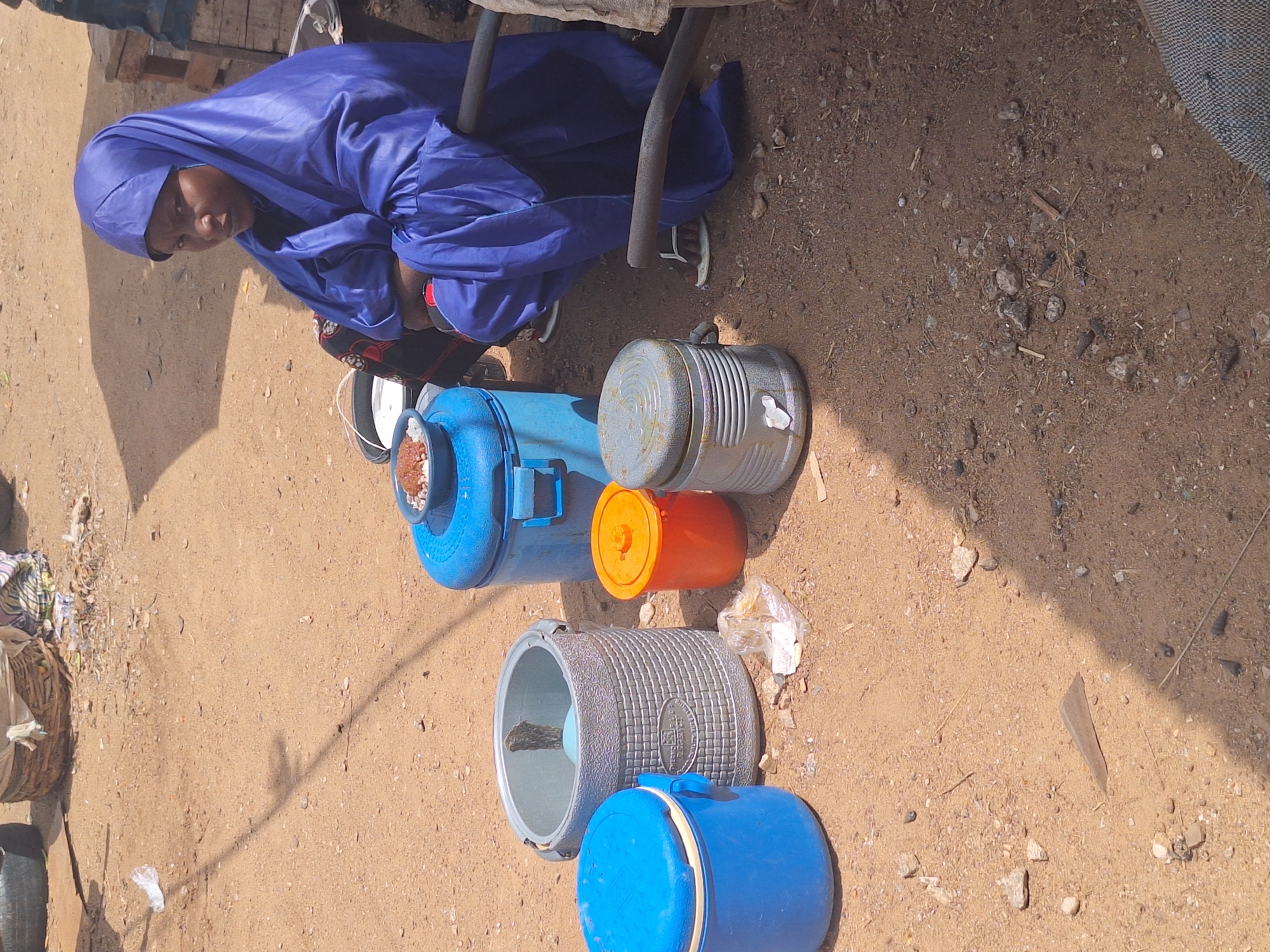 A woman wearing a blue dress and headscarf is seated next to several containers on a dirt surface, possibly in Sabon Tasha, Nigeria. The containers vary in color and size, including blue, orange, and gray. She appears to be selling drinks.
